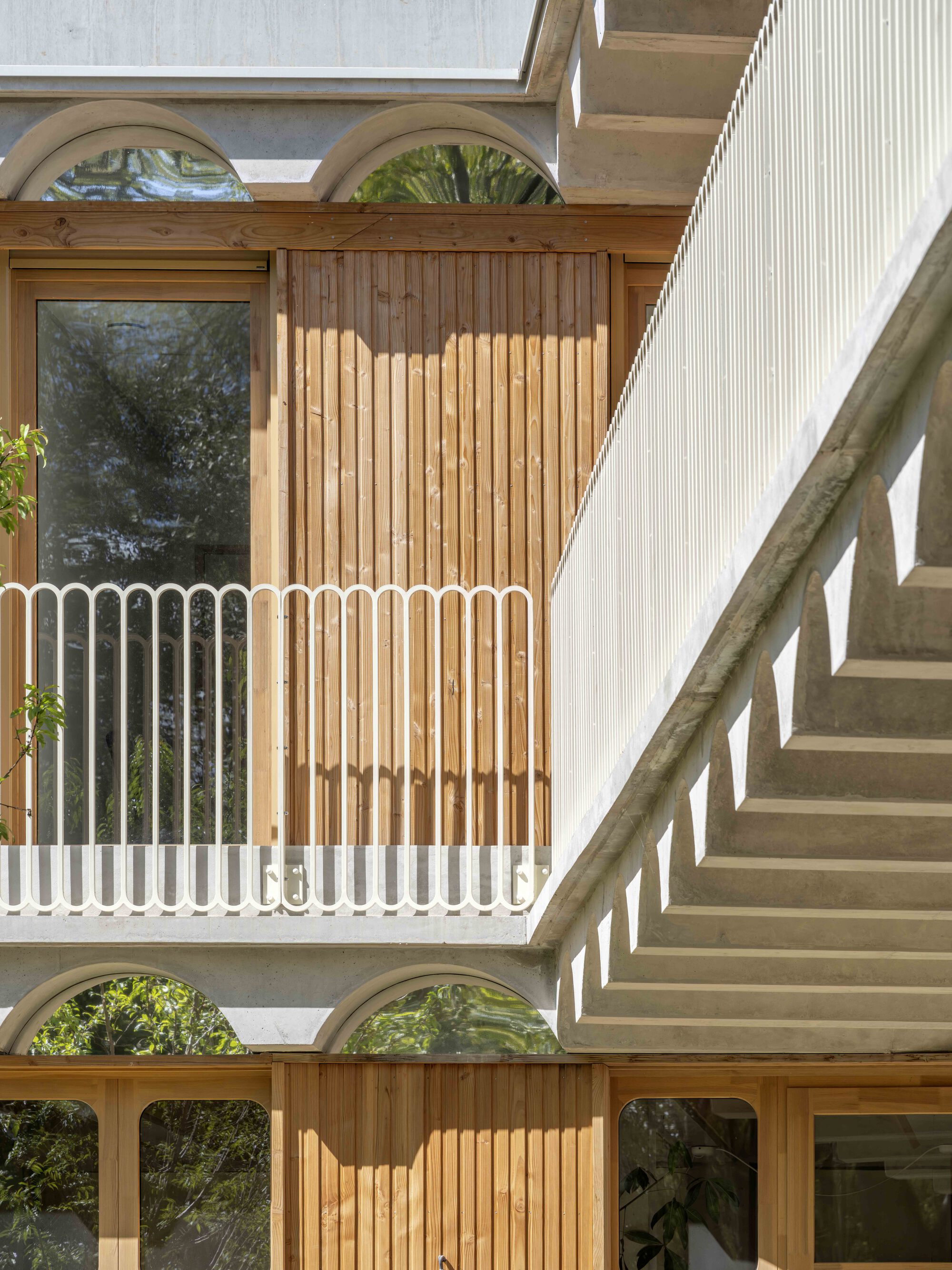 Timber-clad balcony with white arched balustrade beside a concrete staircase soffit, framed by scalloped arches in dappled sunlight