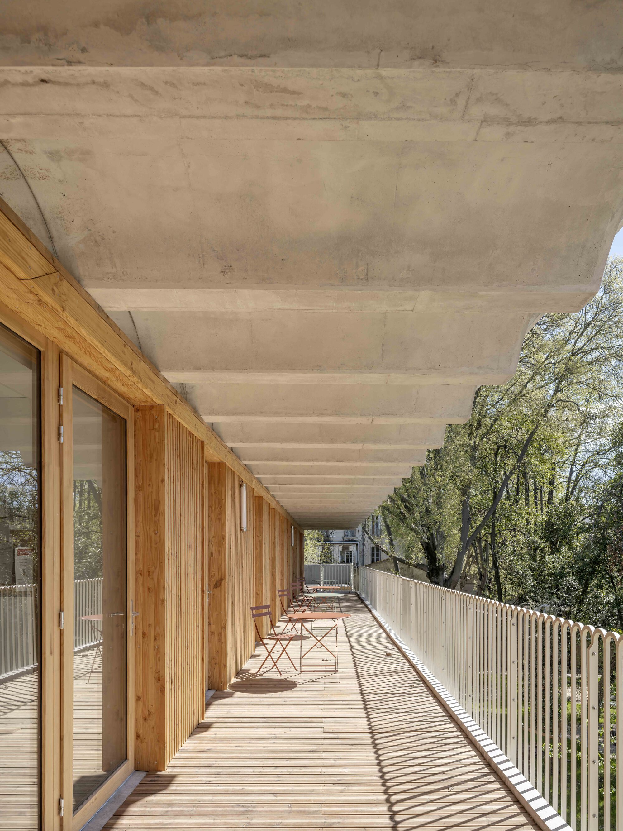 Long upper walkway under a ribbed concrete soffit, with timber doors, bistro chairs and a white railing in afternoon shadow