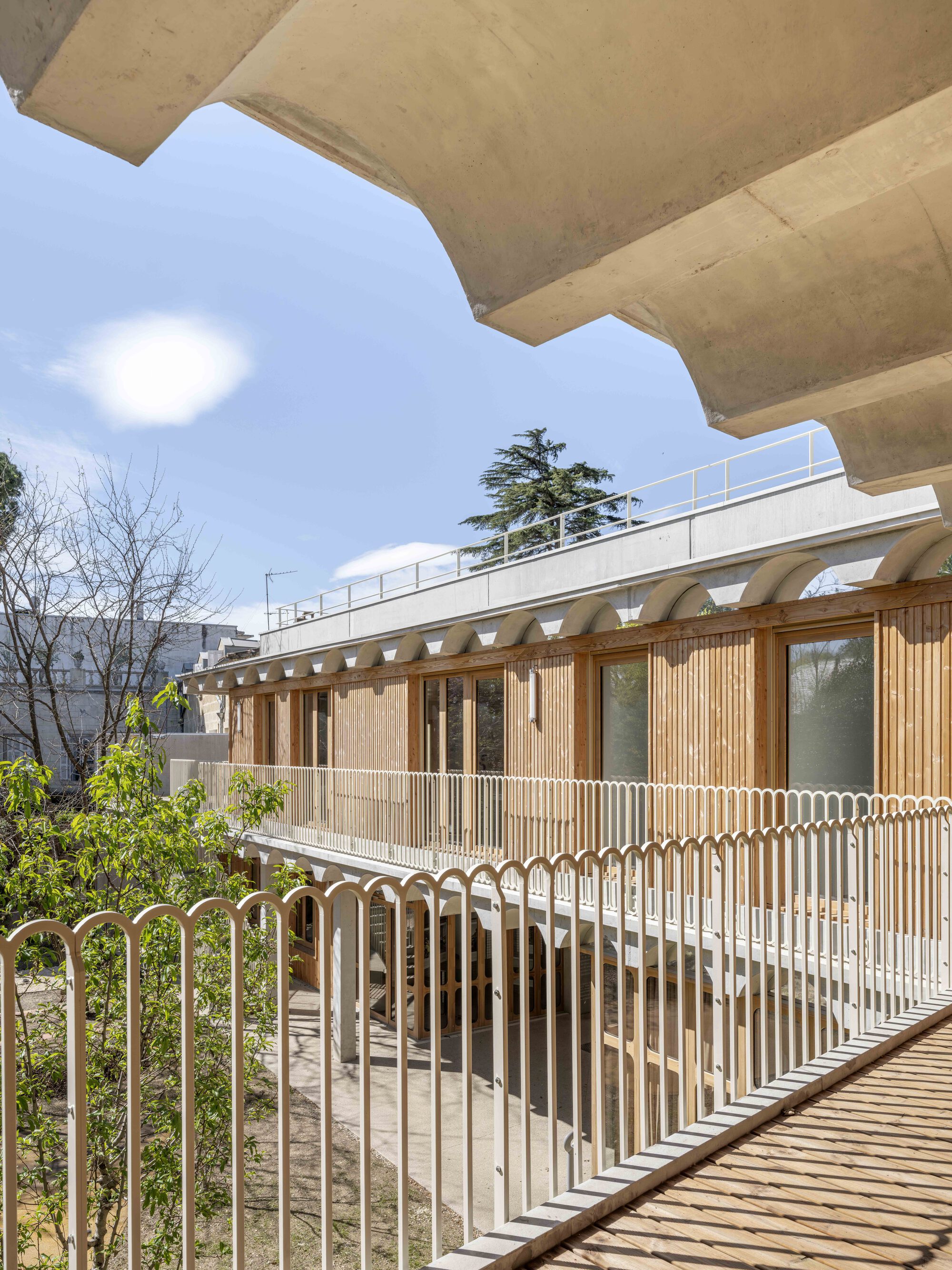 View from a deck past scalloped concrete beams toward a timber-fronted gallery with arched lintels under a blue sky