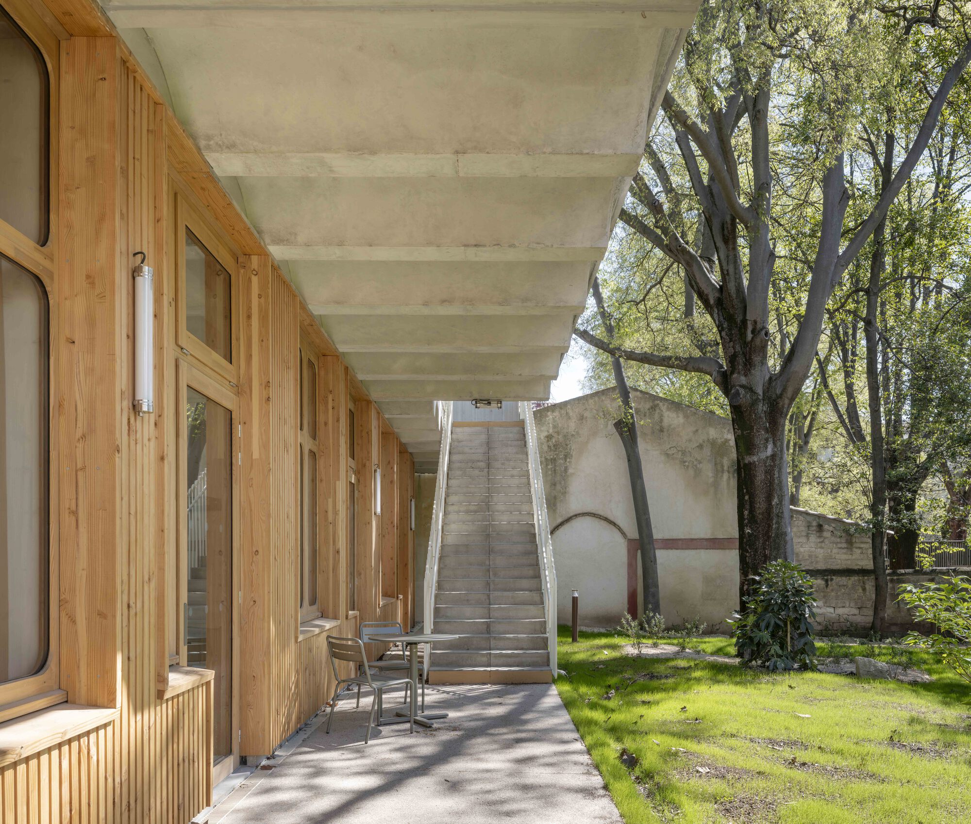 Ground-floor timber arcade under a ribbed concrete ceiling leading to a straight concrete stair beside a lawn and mature tree