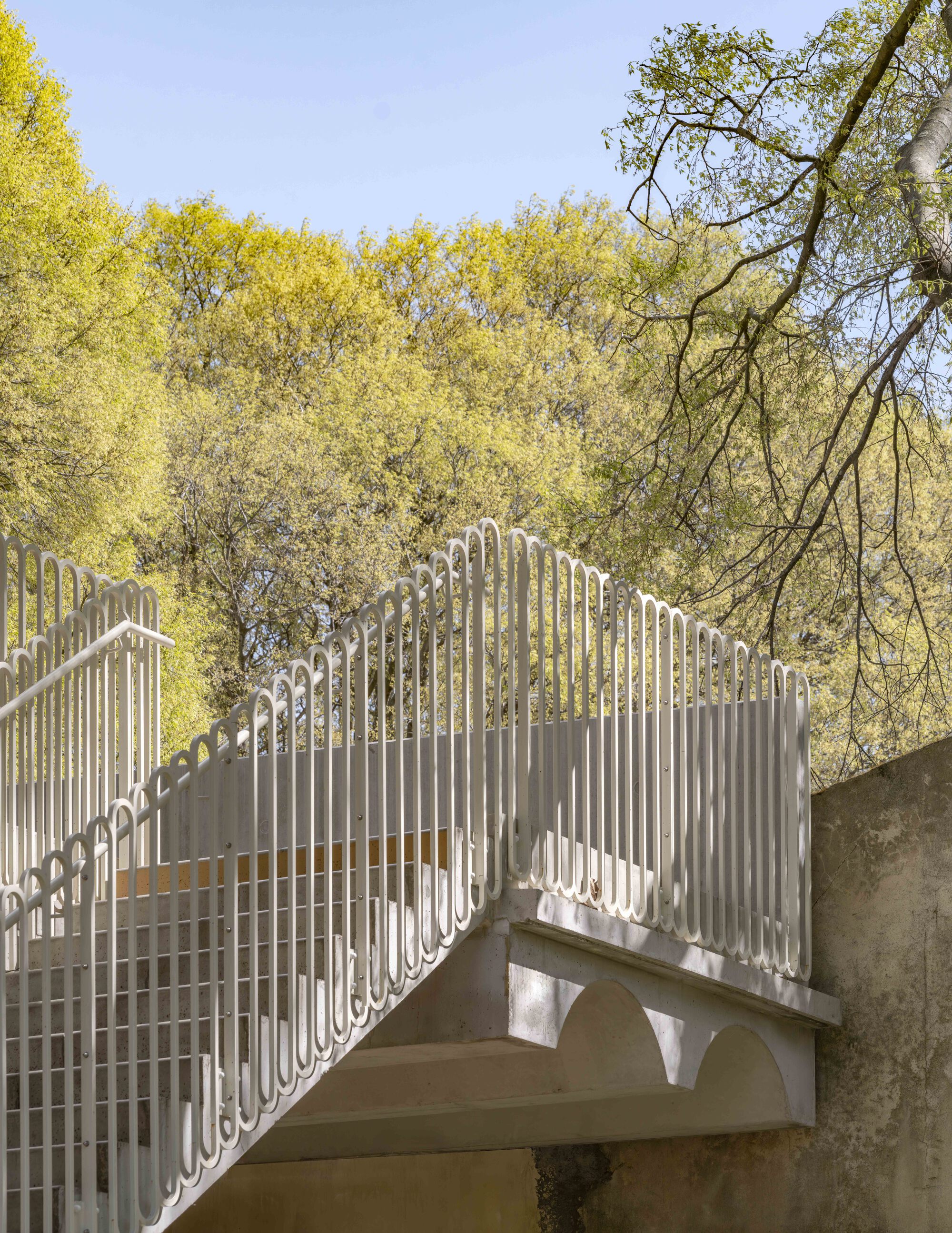 Concrete stair with white looped railing and arched underside, set against fresh spring foliage and a pale blue sky