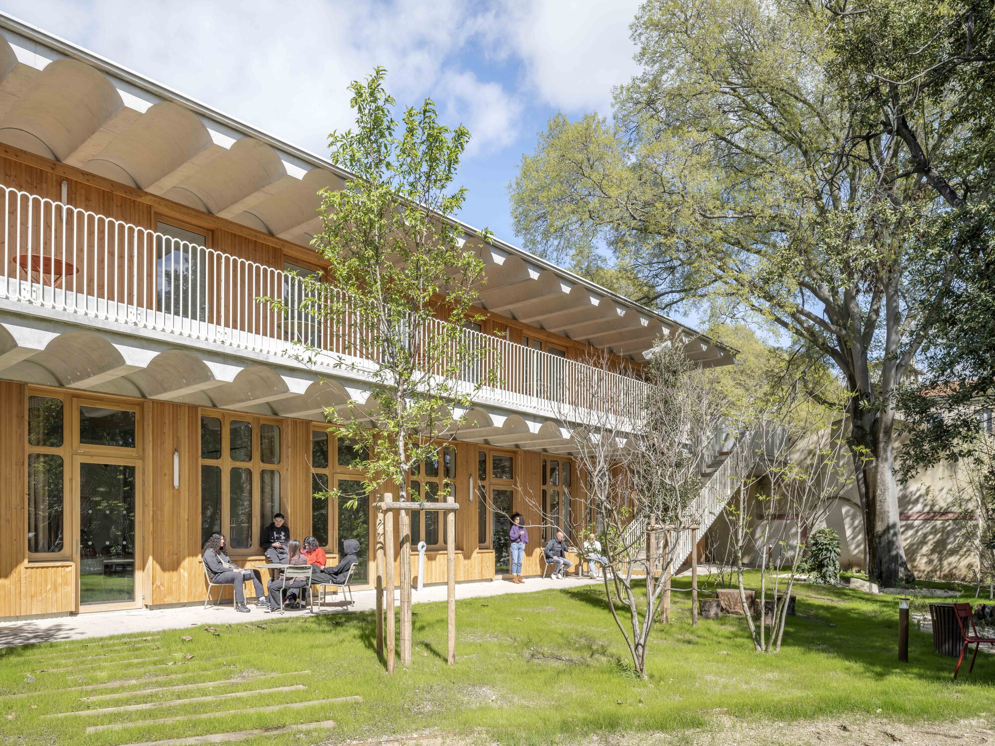 Two-storey timber facade with a scalloped concrete soffit and white balustrade, residents seated outside on a sunny spring lawn