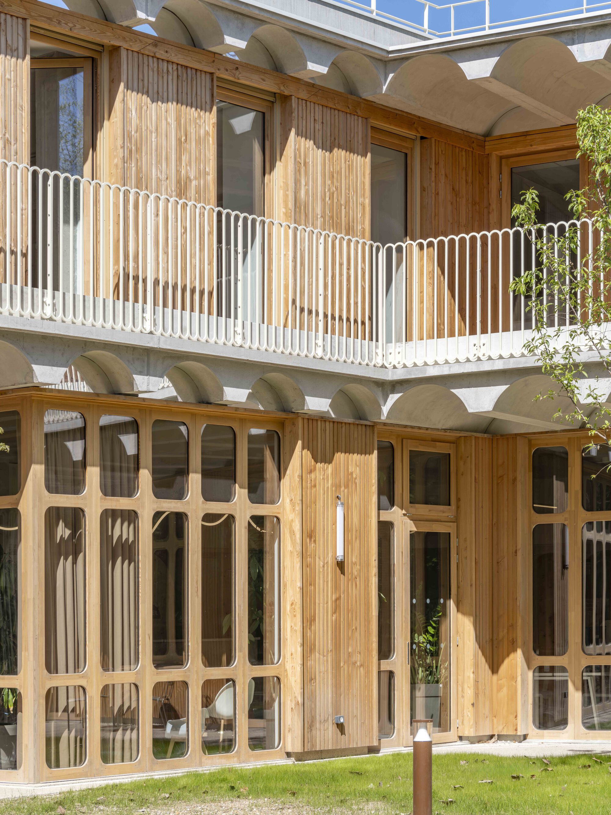 Upper balcony with white railing above arched concrete vaults and tall timber-framed glazing, a leafy branch entering from the right