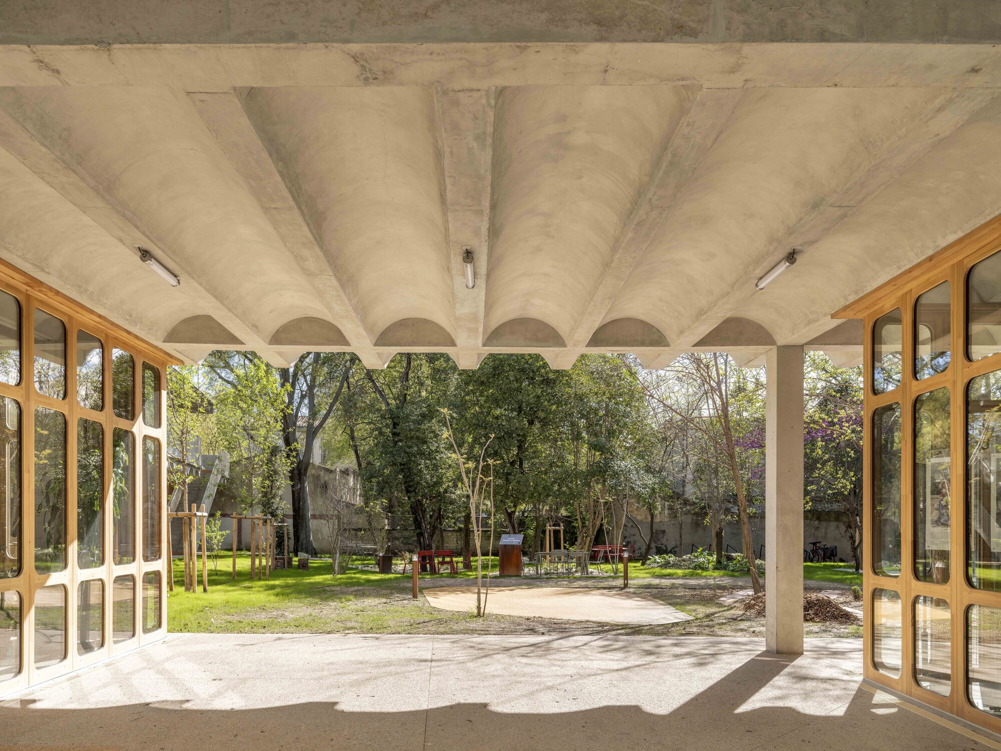 Open ground-floor passage under a barrel-vaulted concrete ceiling, framing a sunlit garden of trees between timber-framed glass walls