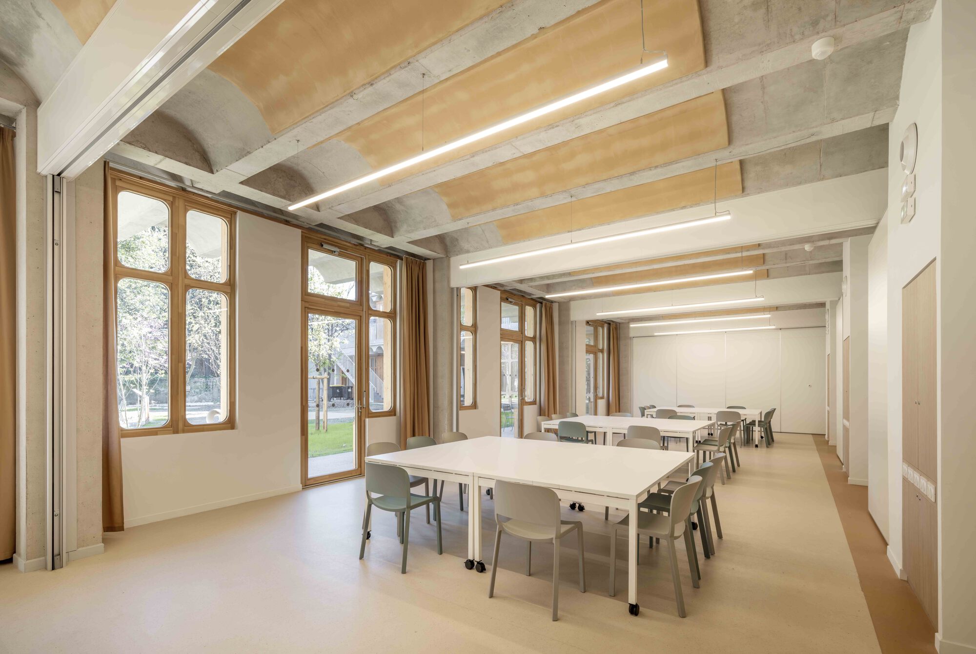 Meeting room with white tables and grey chairs under concrete vaulted ceiling, tall timber-framed windows opening to a sunlit garden