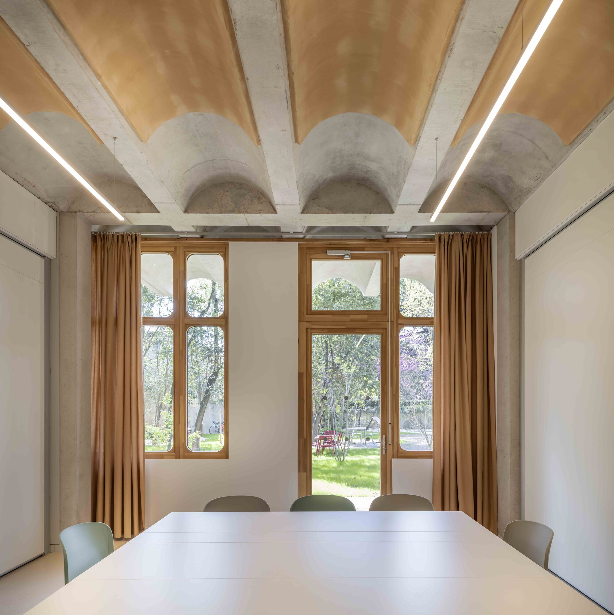 Axial view of a table facing timber-framed windows and a glazed door, with tan curtains and vaulted concrete ceiling above