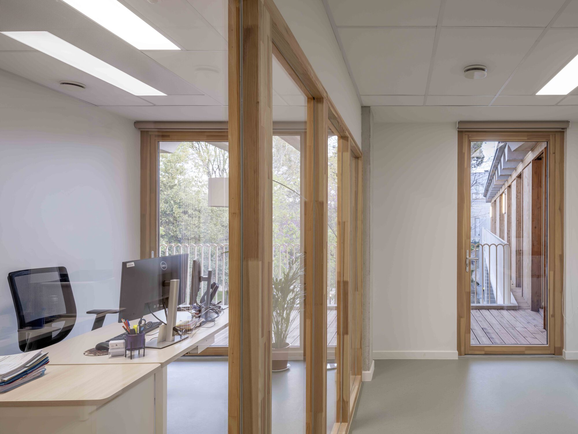 Office desk with monitor beside a timber-framed glass partition, a doorway opening onto a wooden balcony with trees beyond