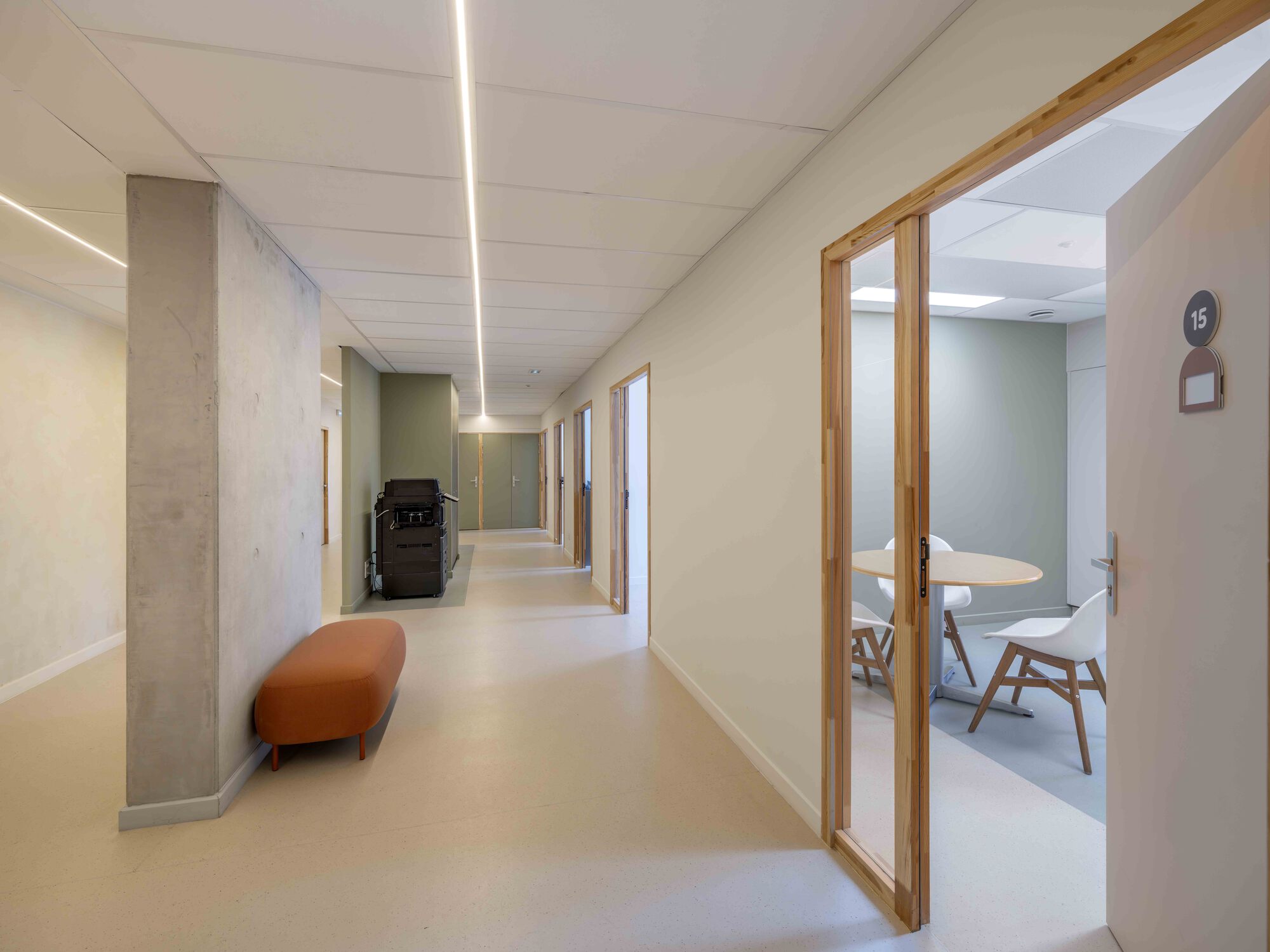 Corridor with a bare concrete column, orange bench and timber-framed glazed door revealing a small round meeting table with white chairs