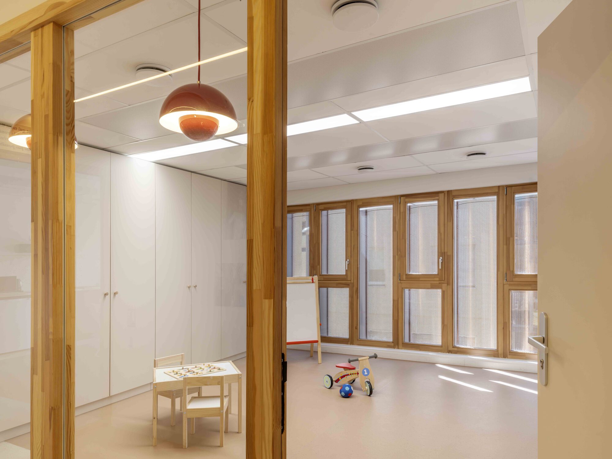 Playroom seen through timber-framed glazing, with a red pendant lamp, white cabinets, a child's tricycle and daylight from tall wood windows