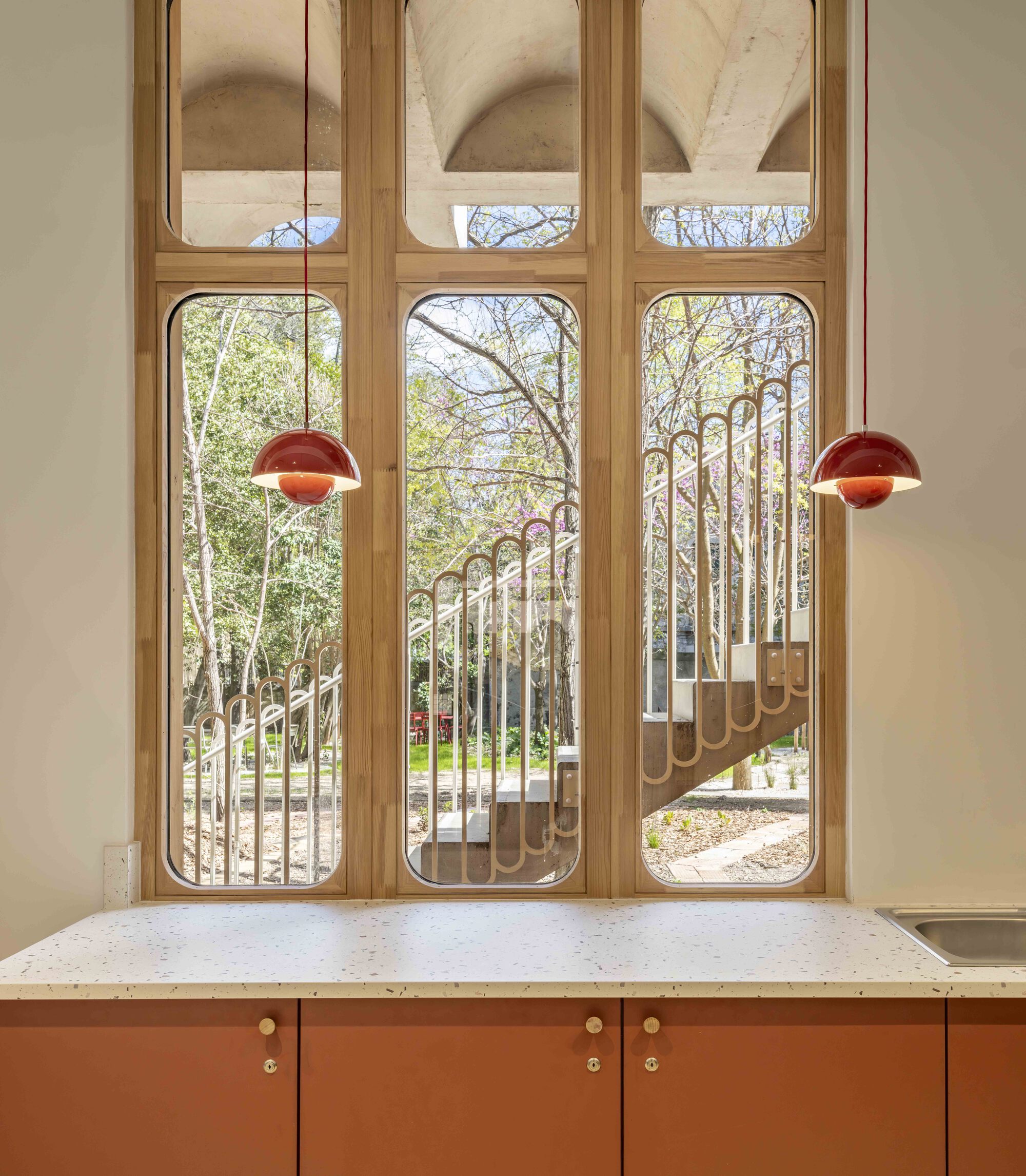 Tall arched timber windows above a terrazzo counter framing a garden stair and bare spring trees, with two red pendant lamps hanging in front