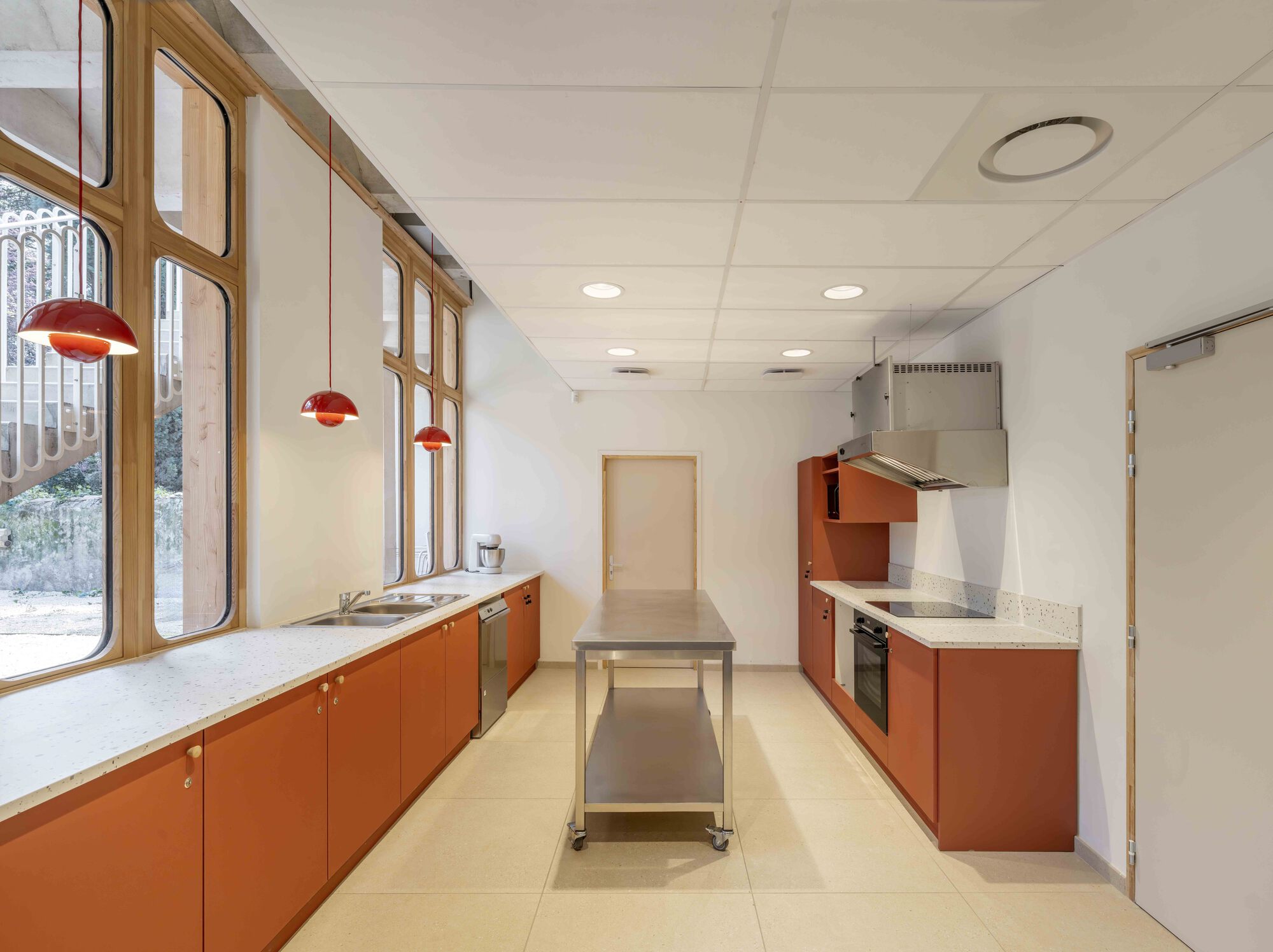 Long kitchen with terracotta cabinets, terrazzo worktops and a steel island, lit by red pendants and a row of arched windows