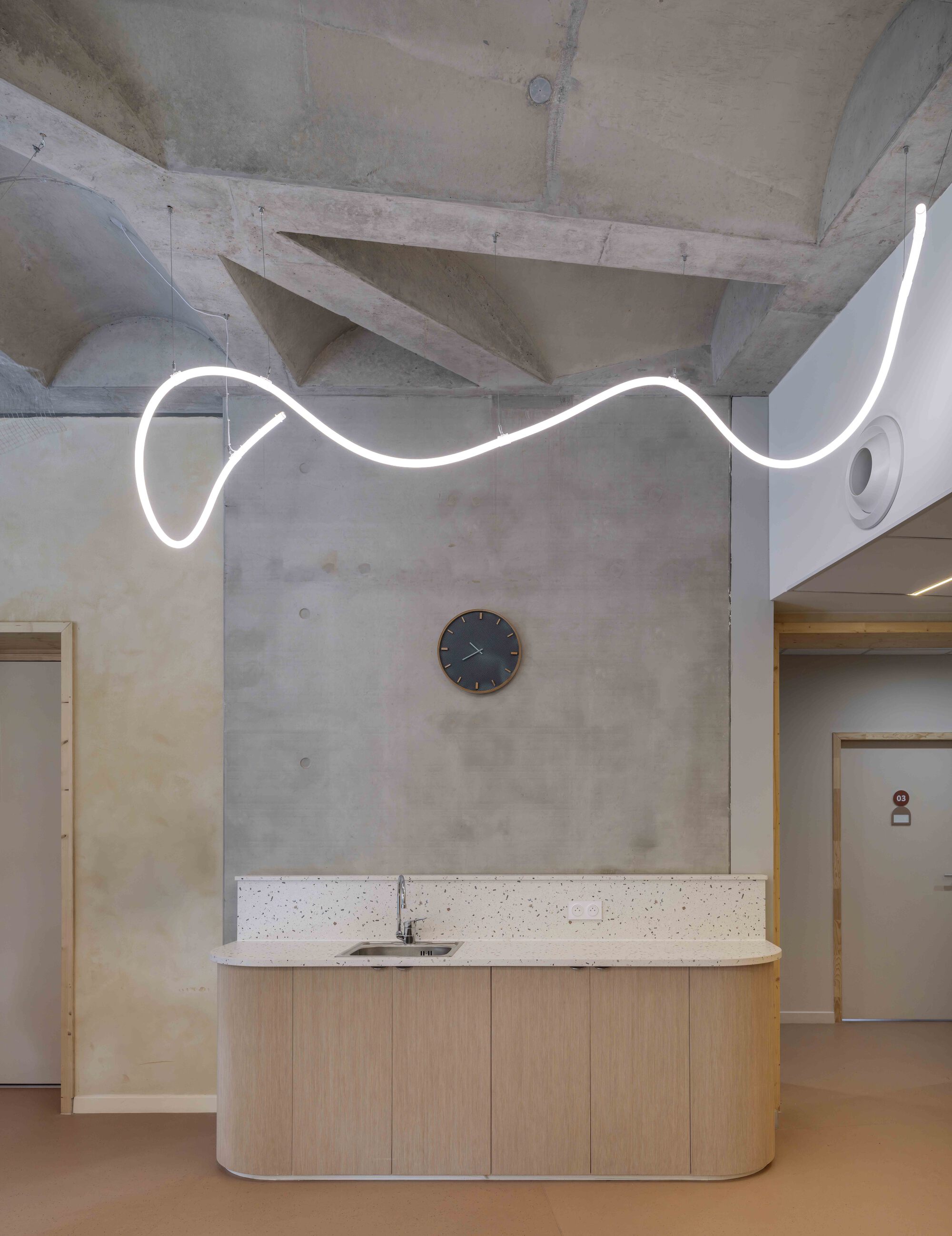 Wavy white light tube hanging below a vaulted concrete ceiling, above a curved timber counter with terrazzo backsplash and a round wall clock
