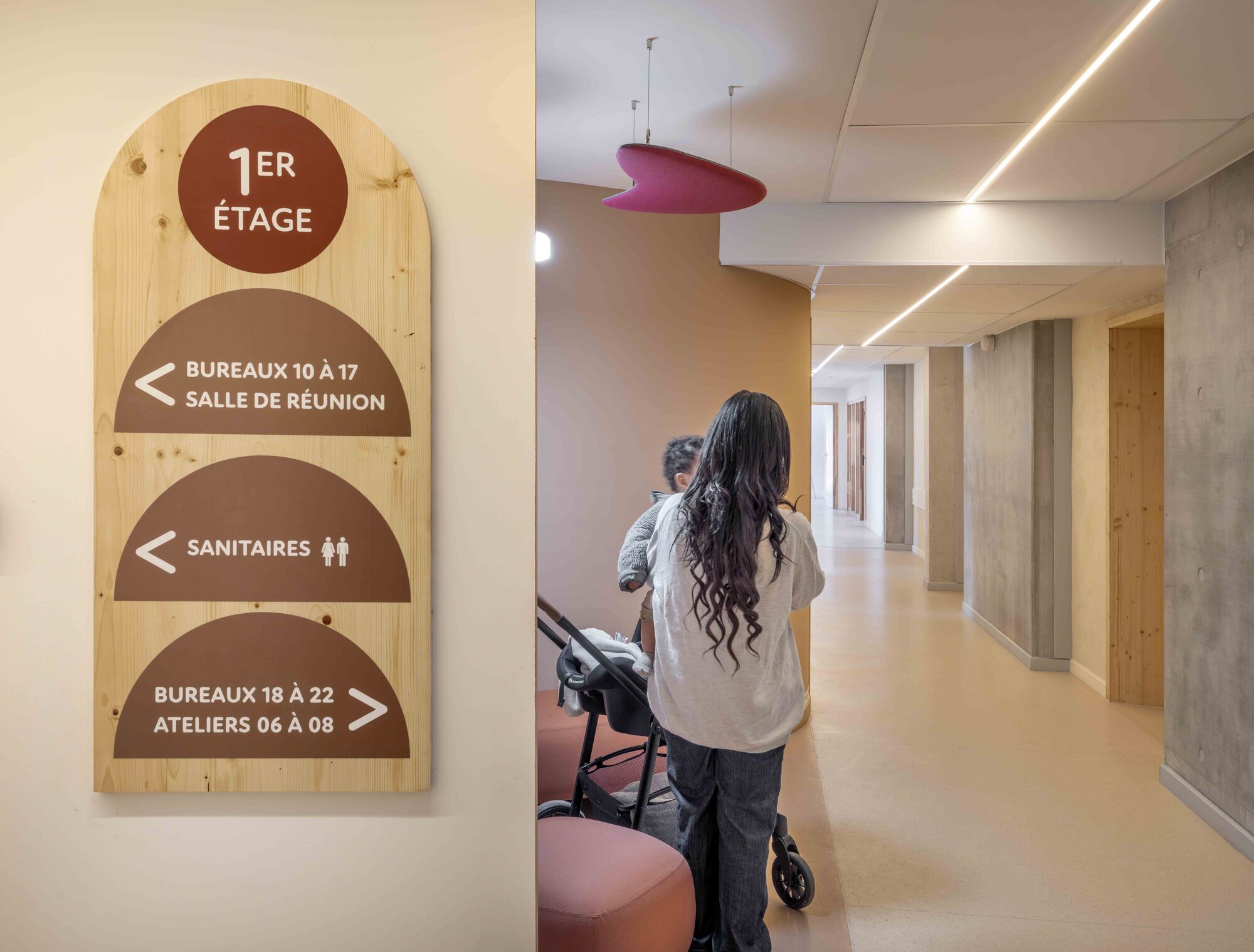 Arched wooden wayfinding sign in French beside a corridor where a woman pushes a stroller past concrete columns and a pink acoustic baffle