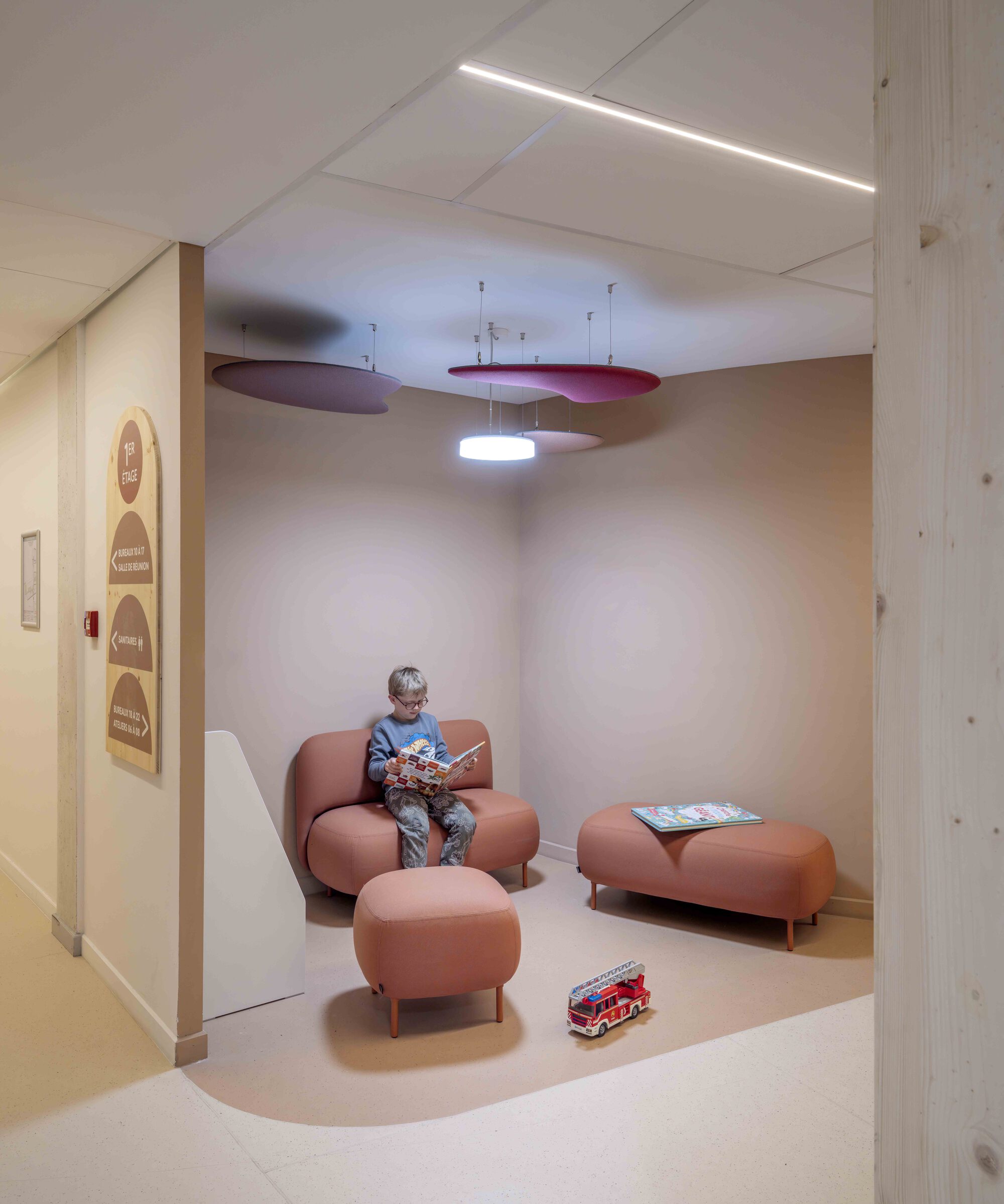 Child reading on a pink sofa in a rounded corridor alcove, with felt ceiling baffles overhead and a toy fire truck on the floor