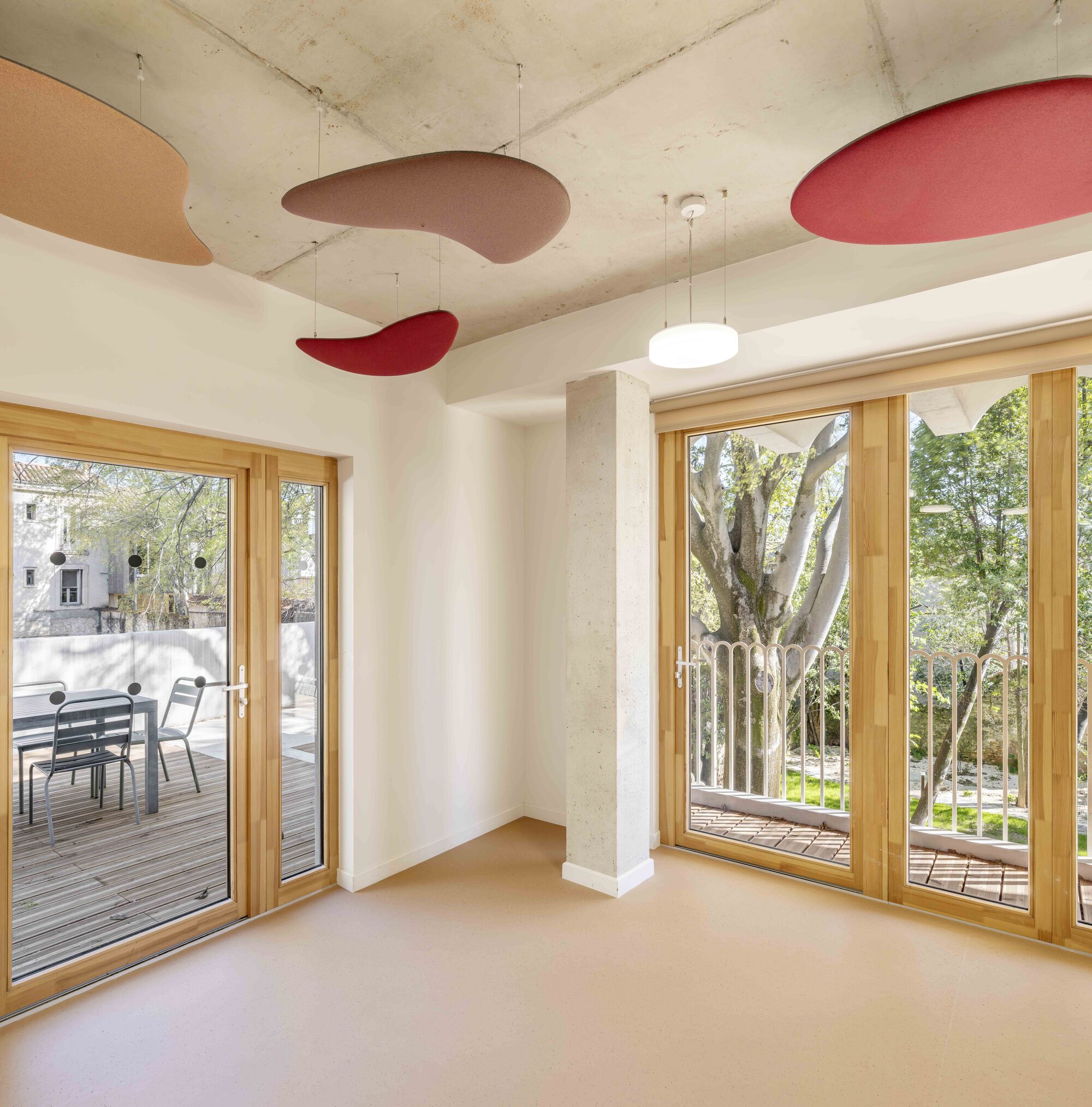 Corner room with timber-framed glazed doors, bare concrete ceiling and suspended red and pink acoustic discs, tree and sunlit balcony outside
