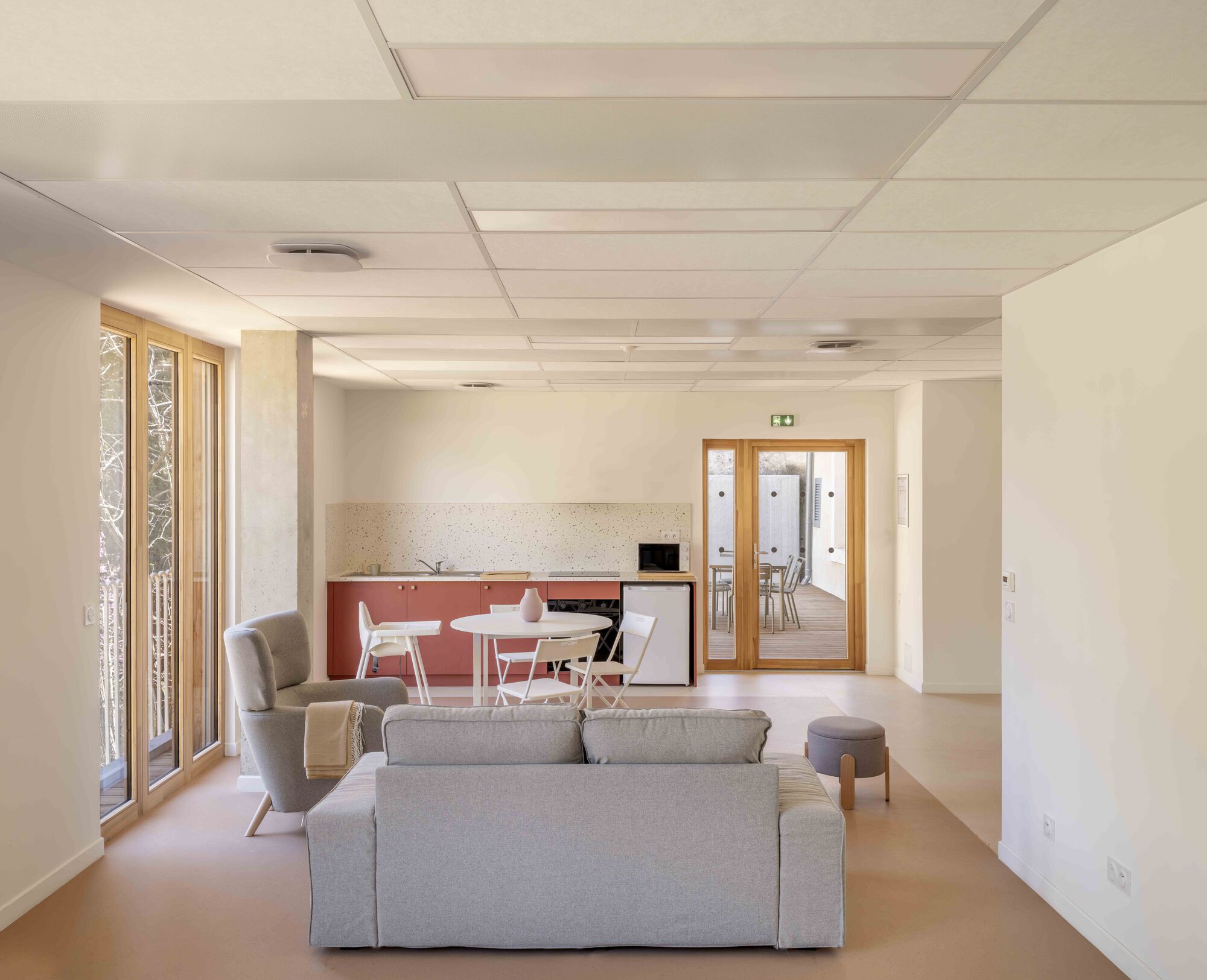 Open common room with grey sofa, terracotta kitchenette and timber-framed doors leading to a deck in soft daylight