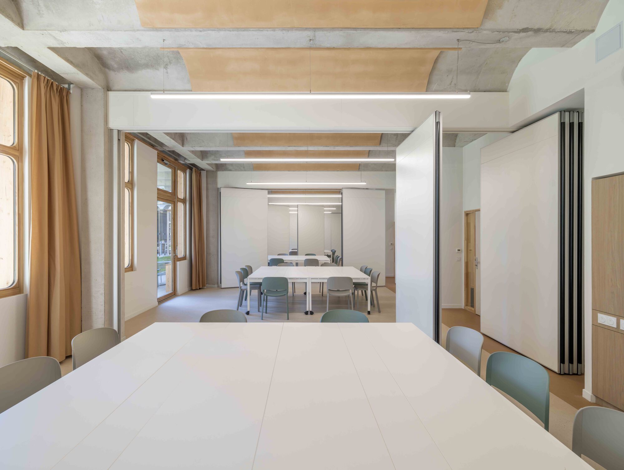Meeting room with white tables and green chairs, sliding partition panels half closed beneath a vaulted concrete and timber ceiling