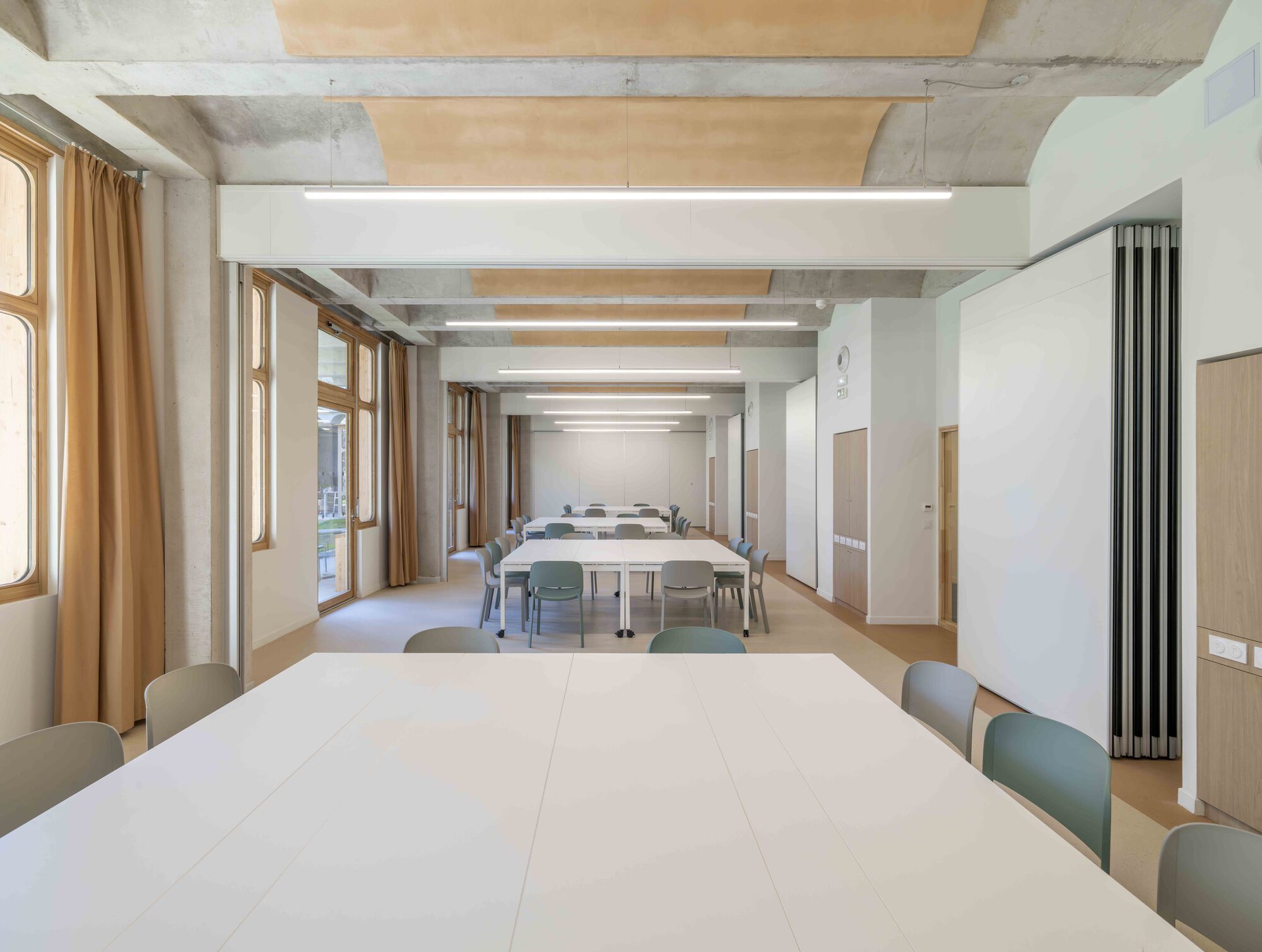 Same meeting room with partitions folded back, long white tables receding under linear light fittings and ochre curtains at the window