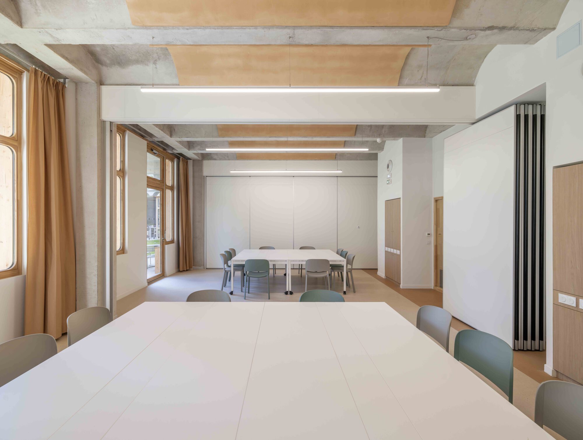 Meeting room closed off by a white folding wall, with one table and grey-green chairs beside ochre curtains and tall windows