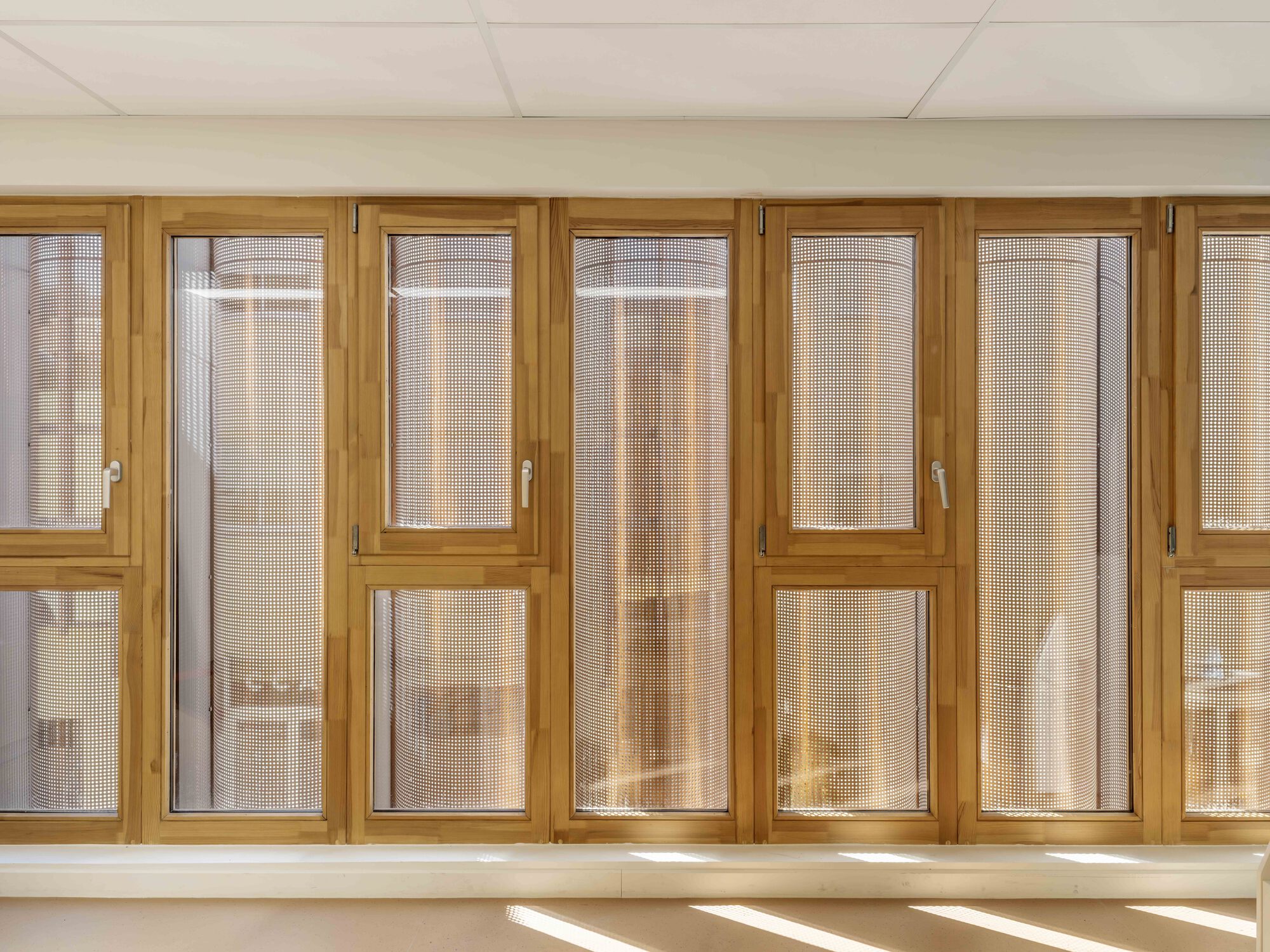 Row of timber-framed windows with perforated screens, sunlight casting bright stripes across the floor below