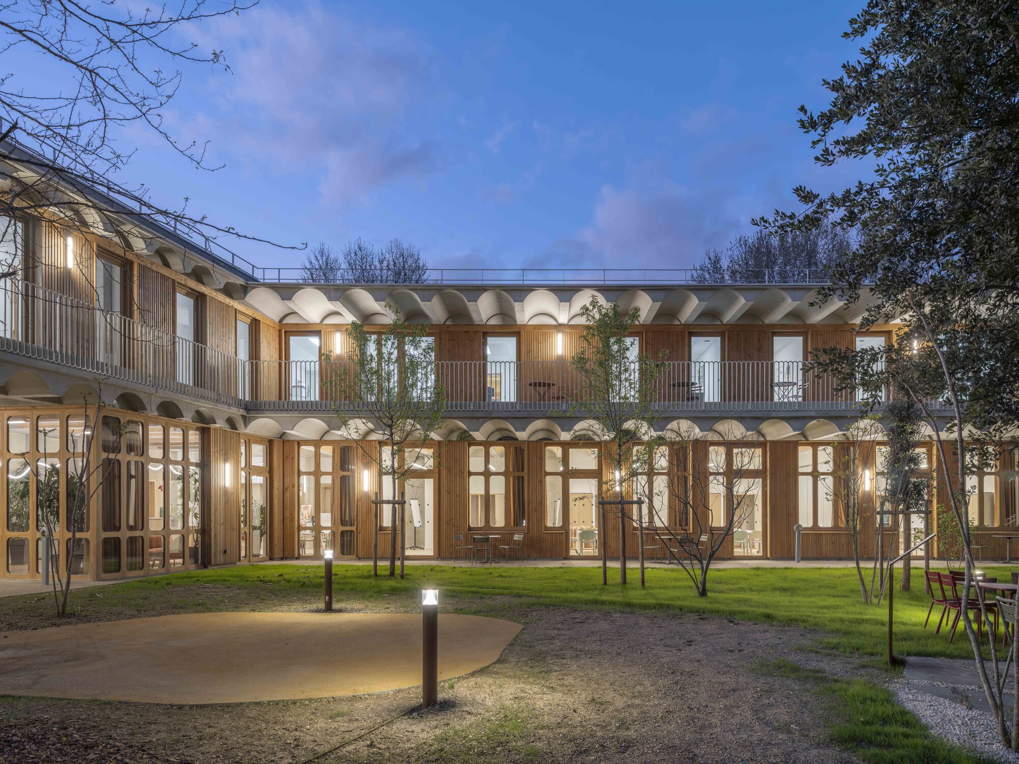 Courtyard at dusk with a lawn, young trees and a two-storey timber facade under scalloped concrete eaves