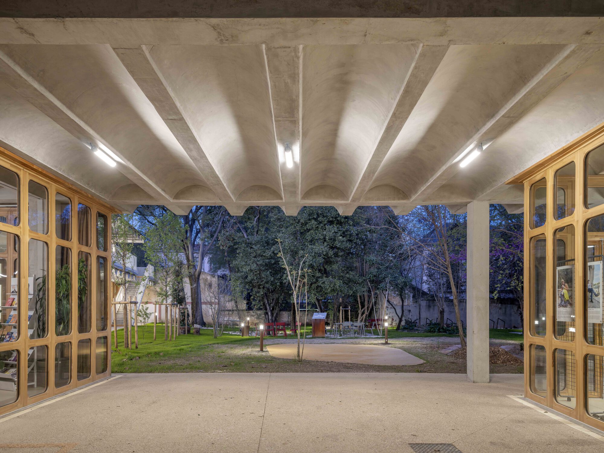 Ribbed concrete vault above an open passage framing a garden, flanked by timber-framed glazed walls at dusk