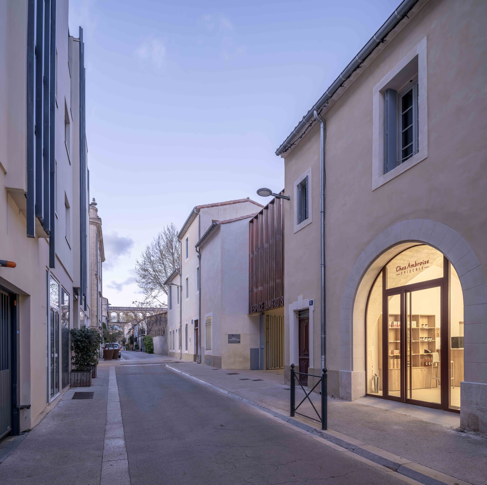 Narrow street at dusk with an arched shopfront glowing on the right and a copper-clad entrance beyond