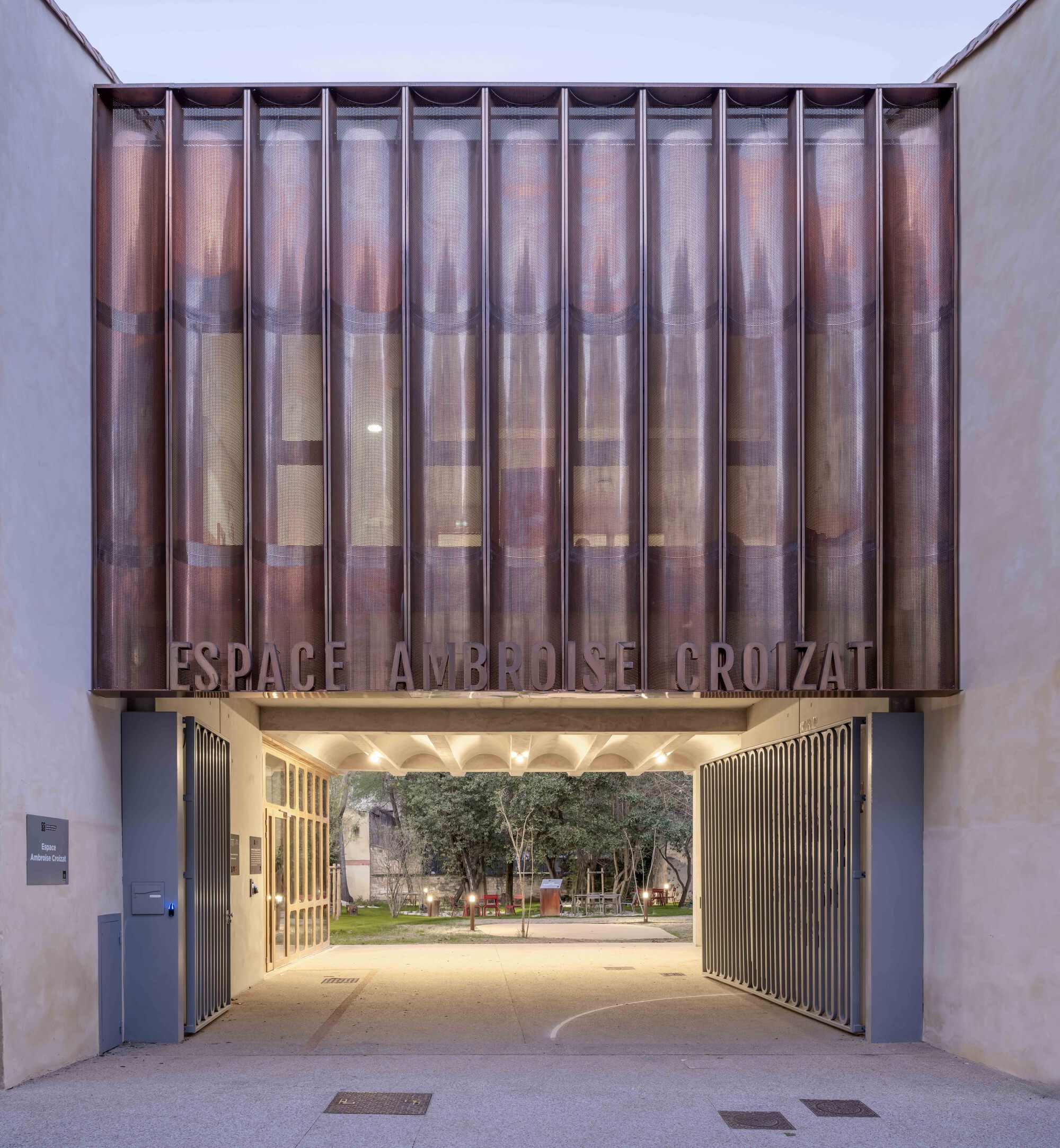 Entrance gateway beneath a pleated copper-toned screen with lettering, sliding metal gates open onto a lit garden at dusk
