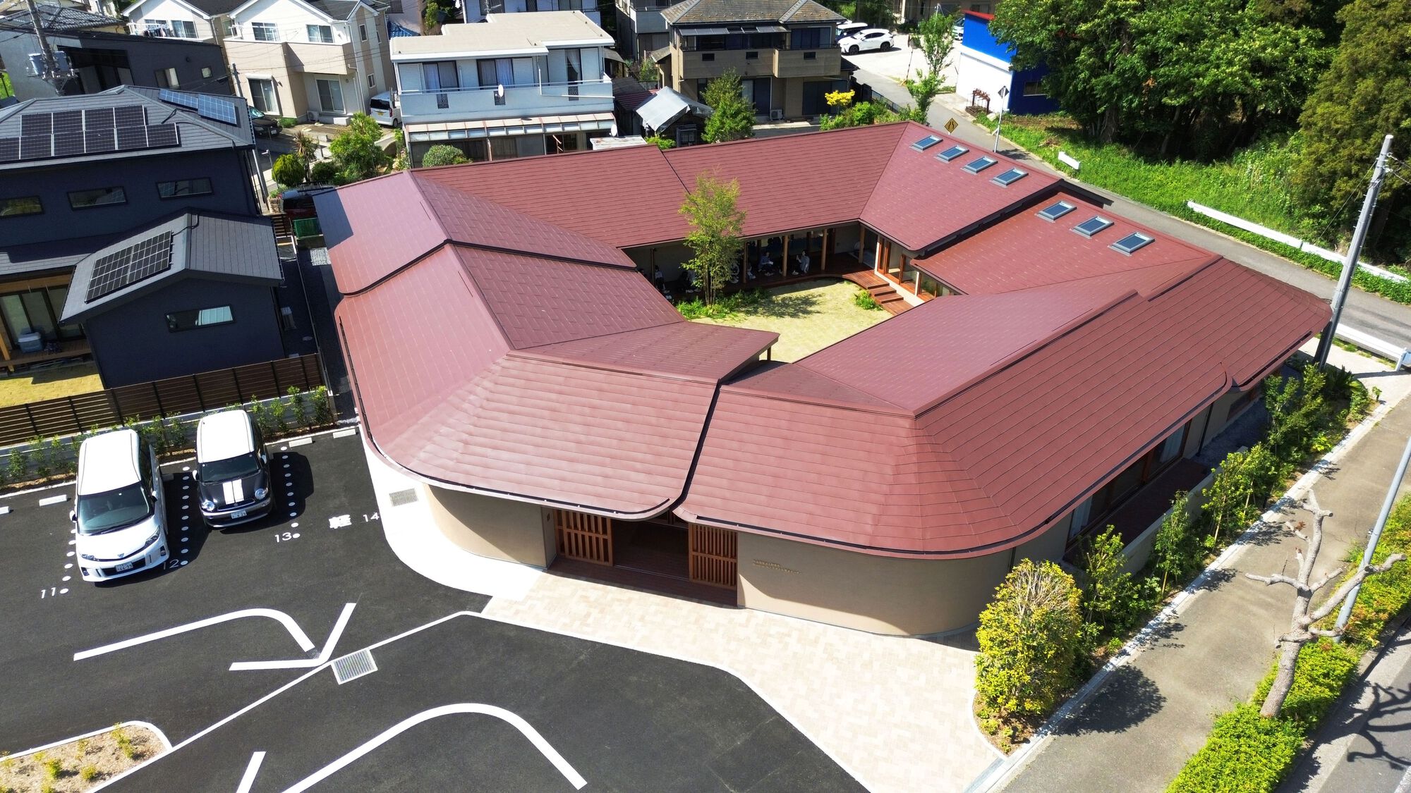 Aerial view of the U-shaped red-brown tiled roof wrapping a grassy courtyard beside a parking lot on a sunny day