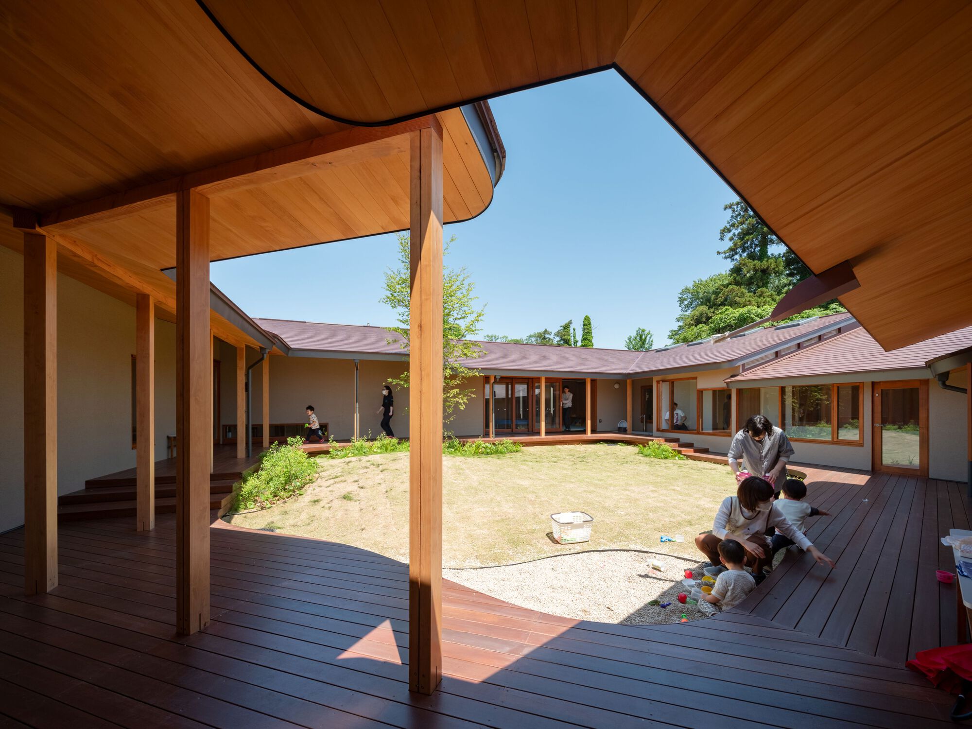 Courtyard seen from a timber deck with slender columns, where a parent and children play on the gravel edge in sunlight