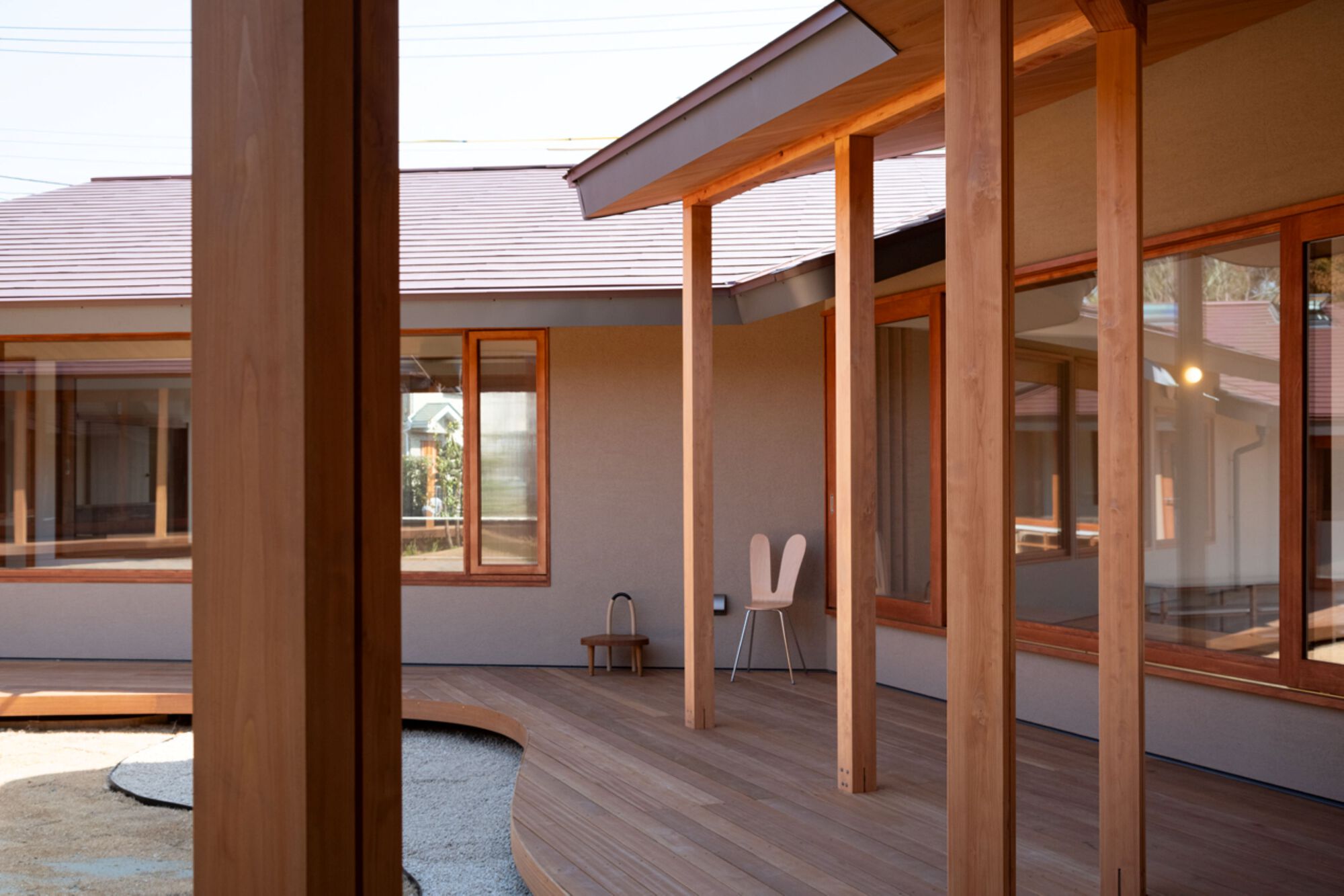 Deck edge curving along glazed walls with timber window frames, a small bunny-eared chair and stool in soft afternoon light