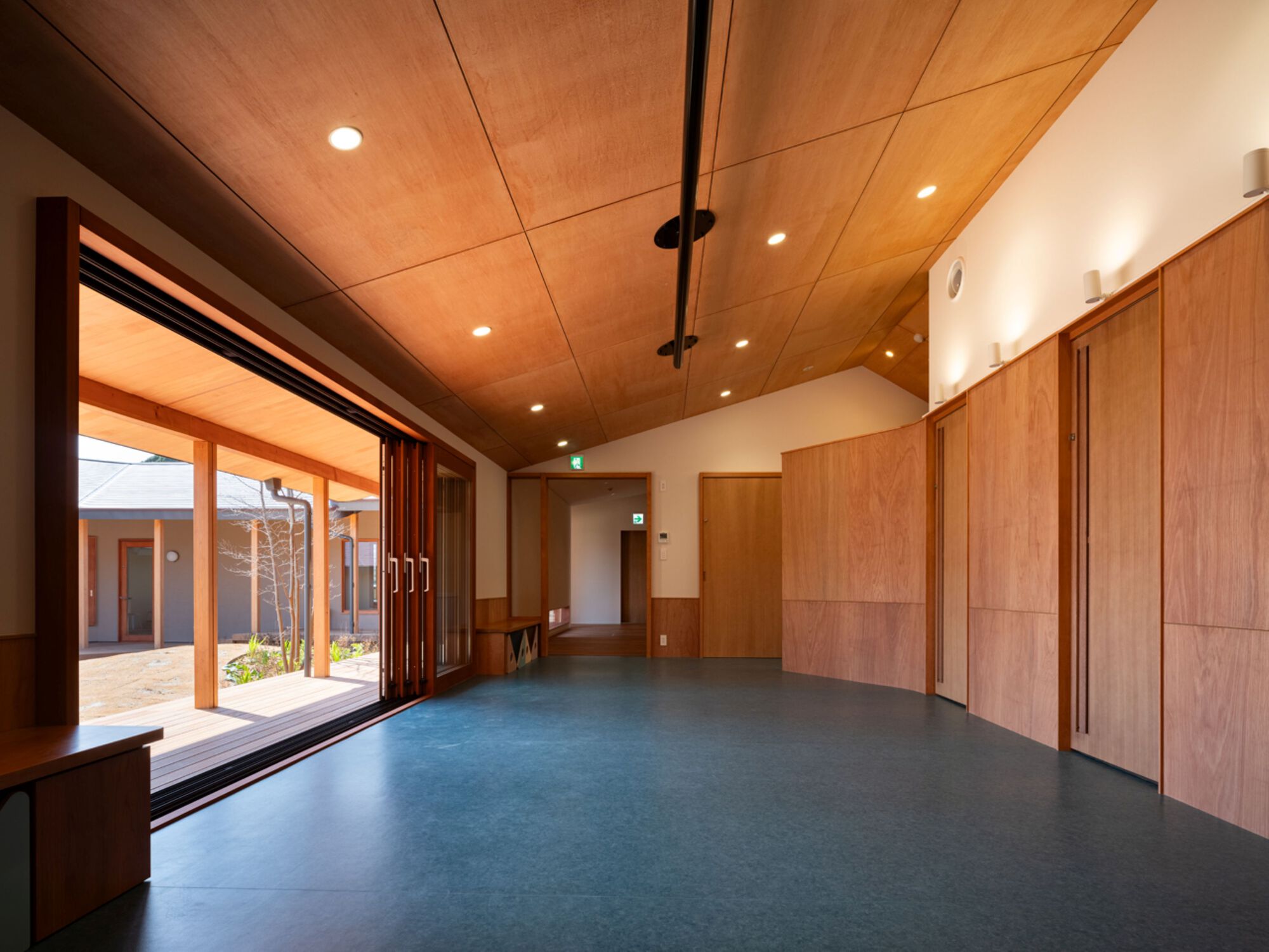 Interior room with sloping plywood ceiling, curved wood wall panels and a sliding glass door opening to the courtyard deck
