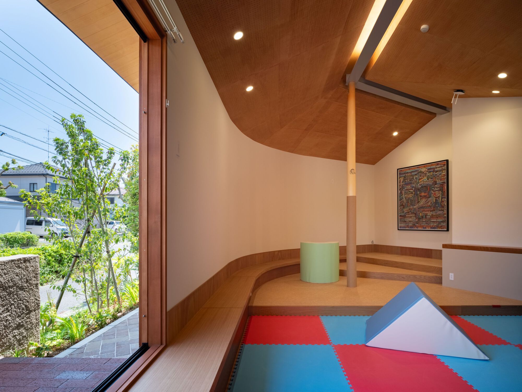 Play room with stepped wooden platform, curved plaster wall, foam floor tiles and a tall window onto a planted street edge