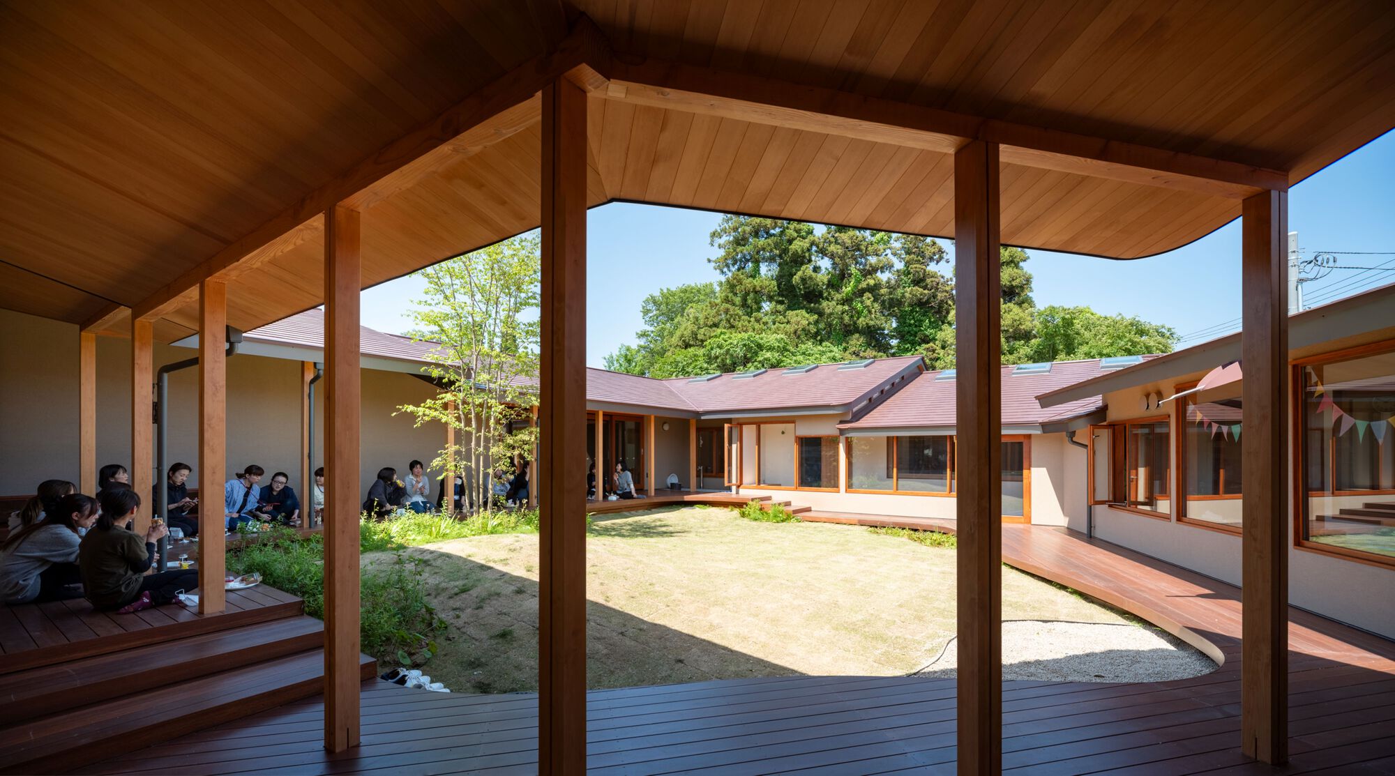 View from a timber-ceilinged veranda across a grassy courtyard, with people seated on deck steps beneath a leafy tree