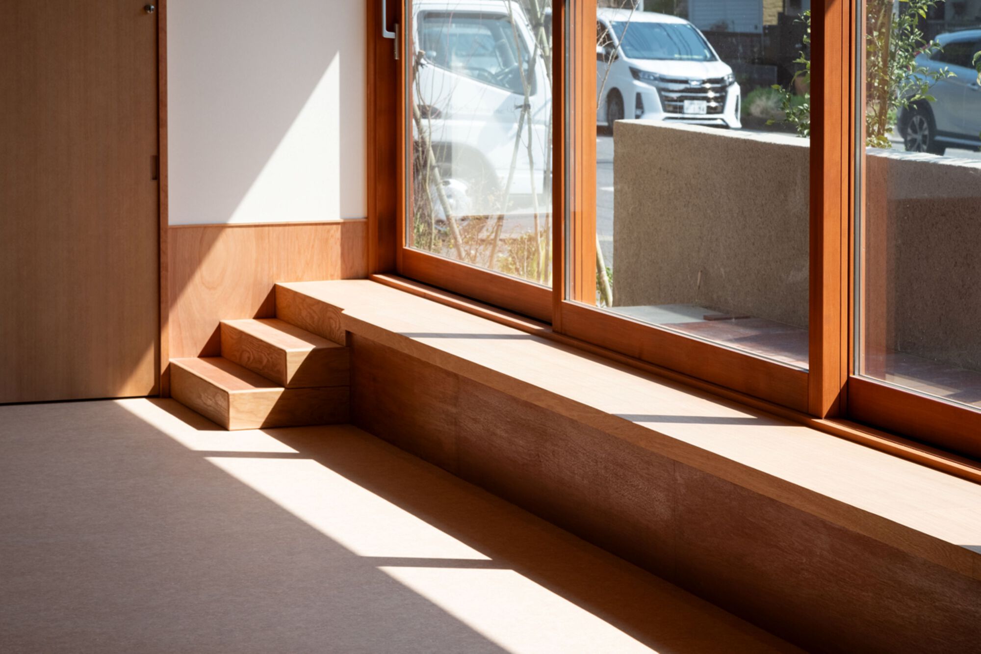 Timber window seat and three small steps beside floor-to-ceiling wood-framed glazing, with sunlight cutting across the carpeted floor