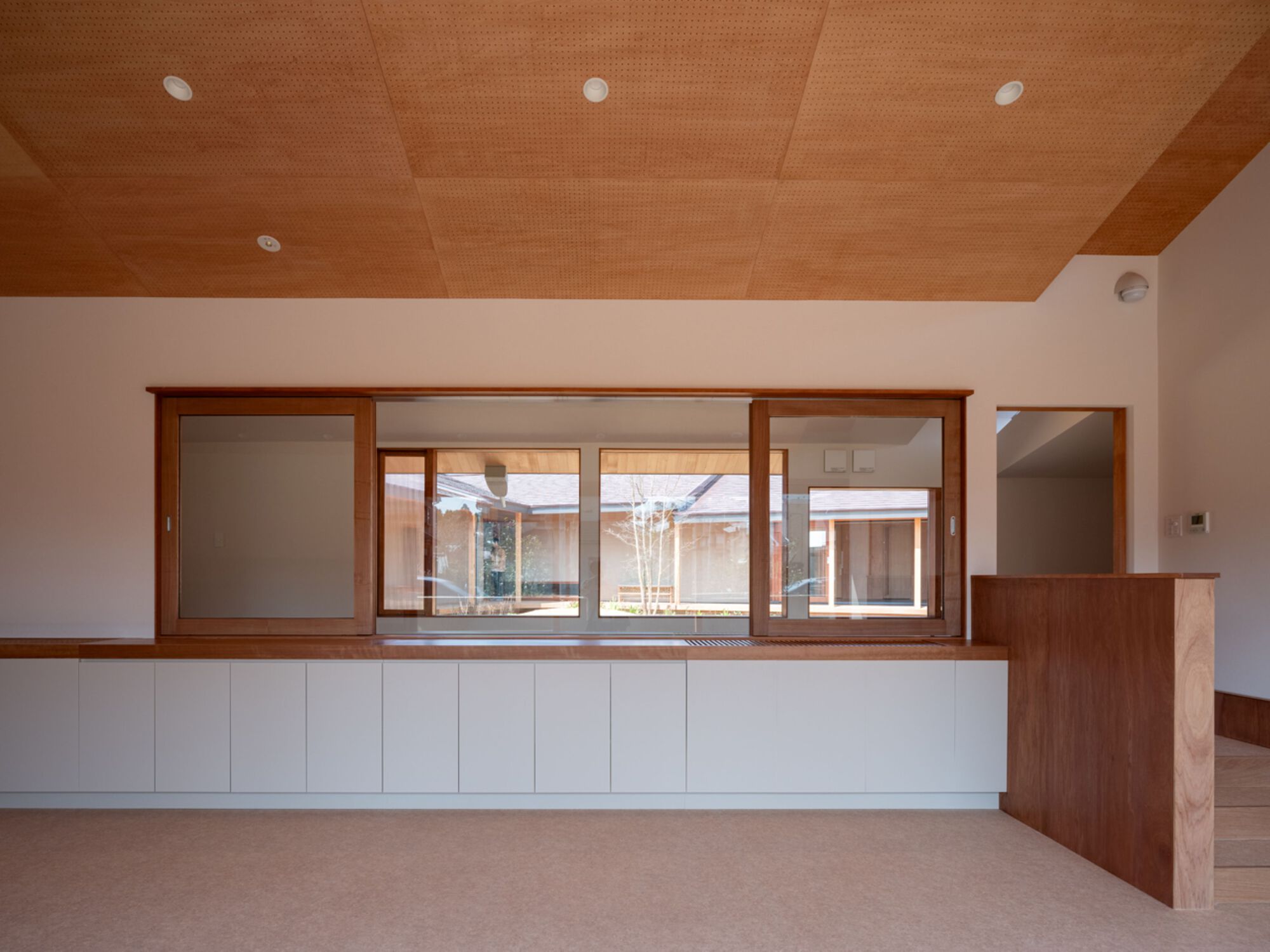 Interior with white built-in cabinets under a sliding timber-framed window opening onto a courtyard, below a perforated plywood ceiling