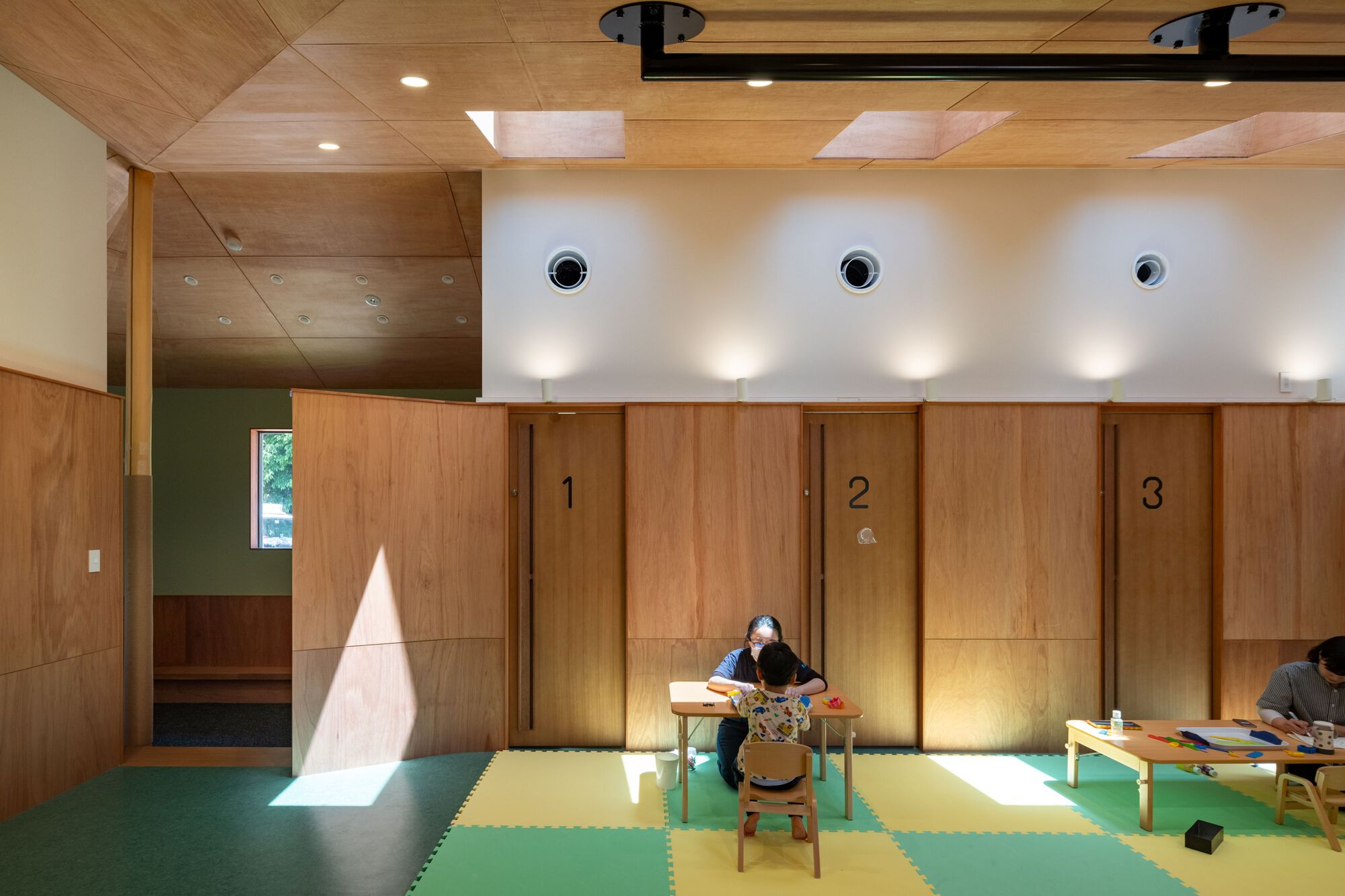Children drawing at low tables on a checkered green and yellow mat in front of numbered plywood doors, with sunlight falling across the wall