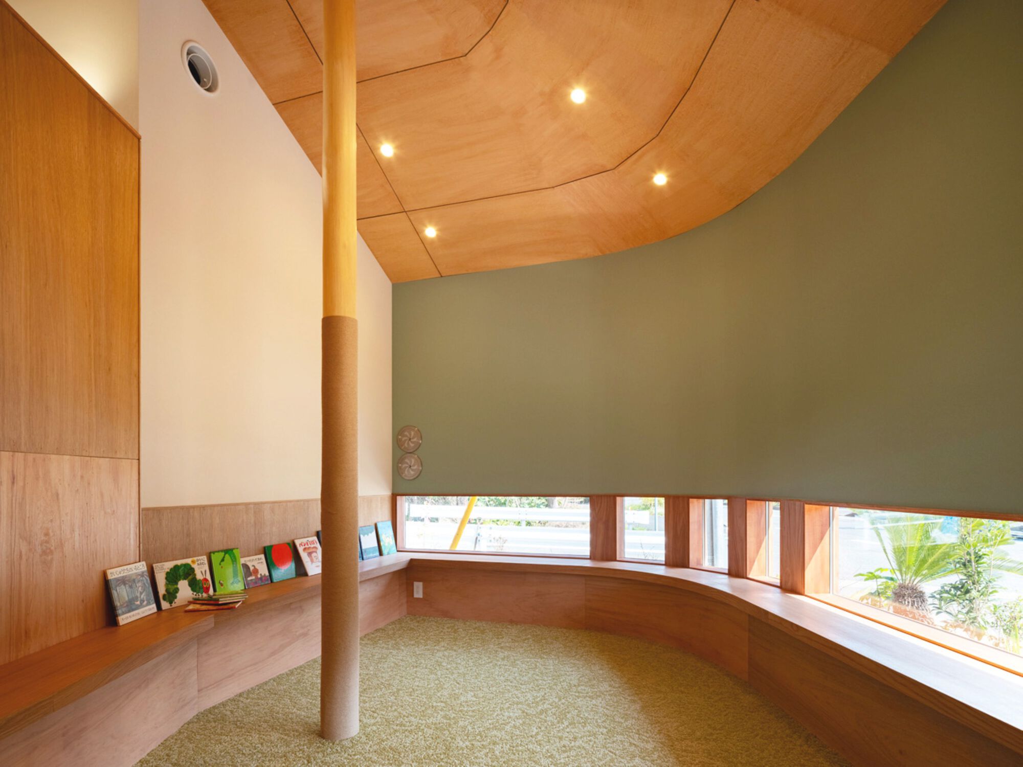 Curved reading nook with a wrapped timber column, a continuous window bench, picture books on a ledge and a sage green wall
