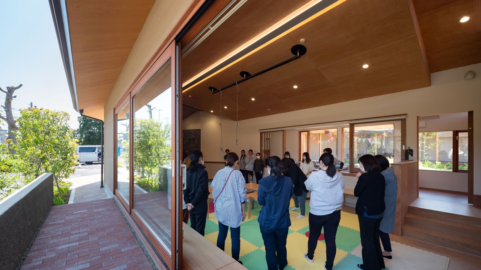 Open sliding glass wall of a timber-ceilinged room where a group of masked adults stand on colorful foam floor mats beside a brick-paved walkway
