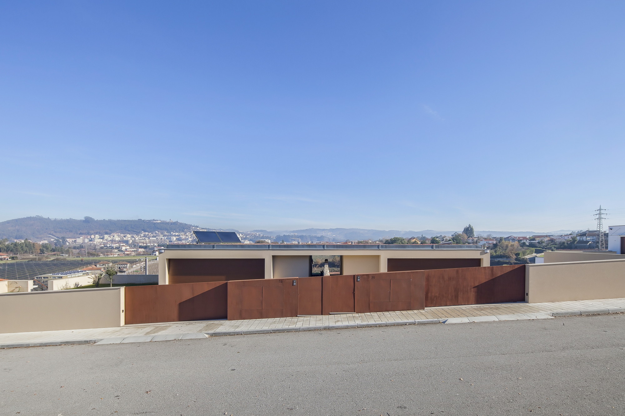 Street-side weathering steel gate and rendered boundary wall, with a low house behind and a town spread across the valley under clear sky