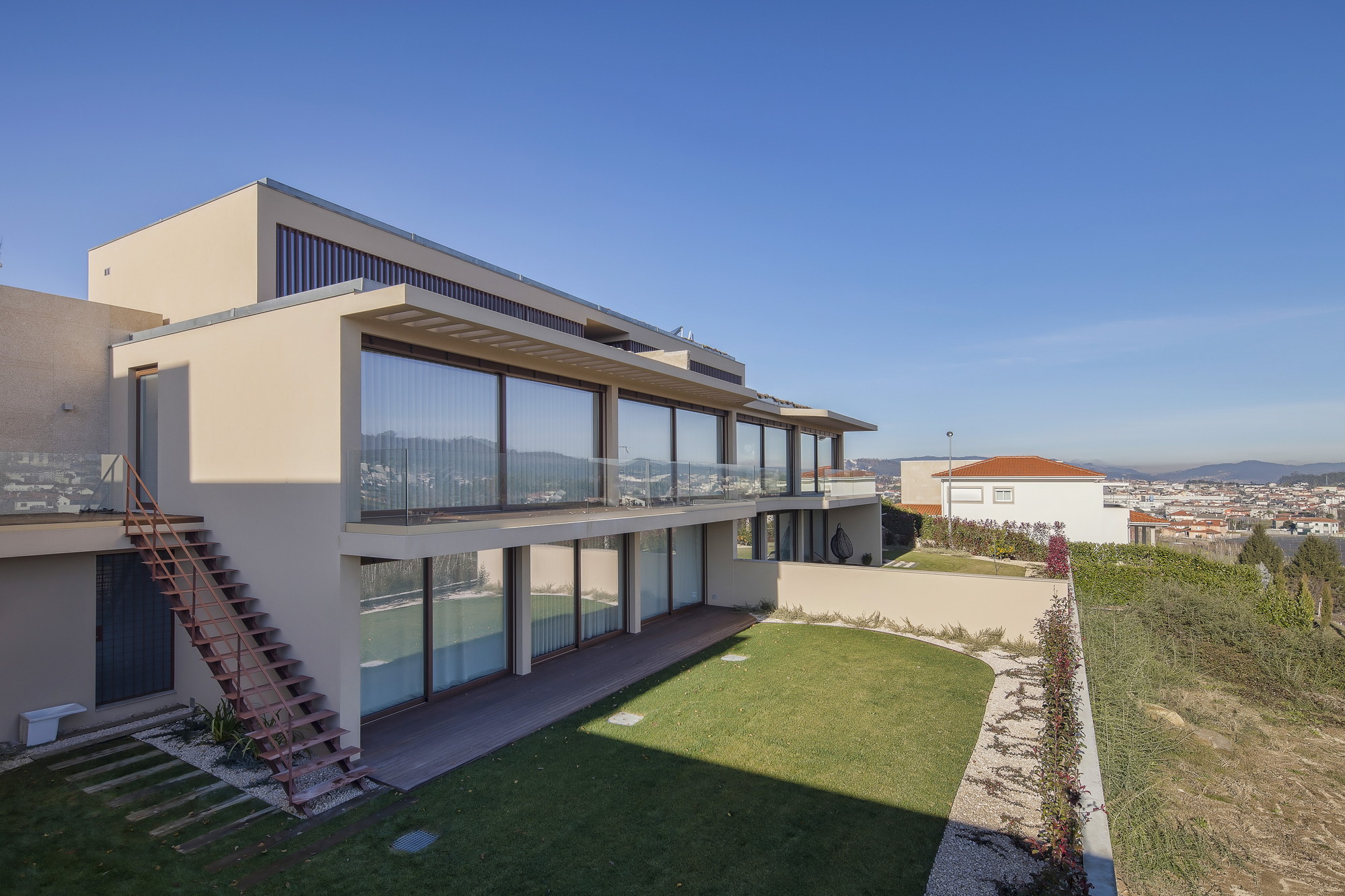 Stepped garden facade with floor-to-ceiling glazing, a rust-colored steel staircase and a lawn in low afternoon sun