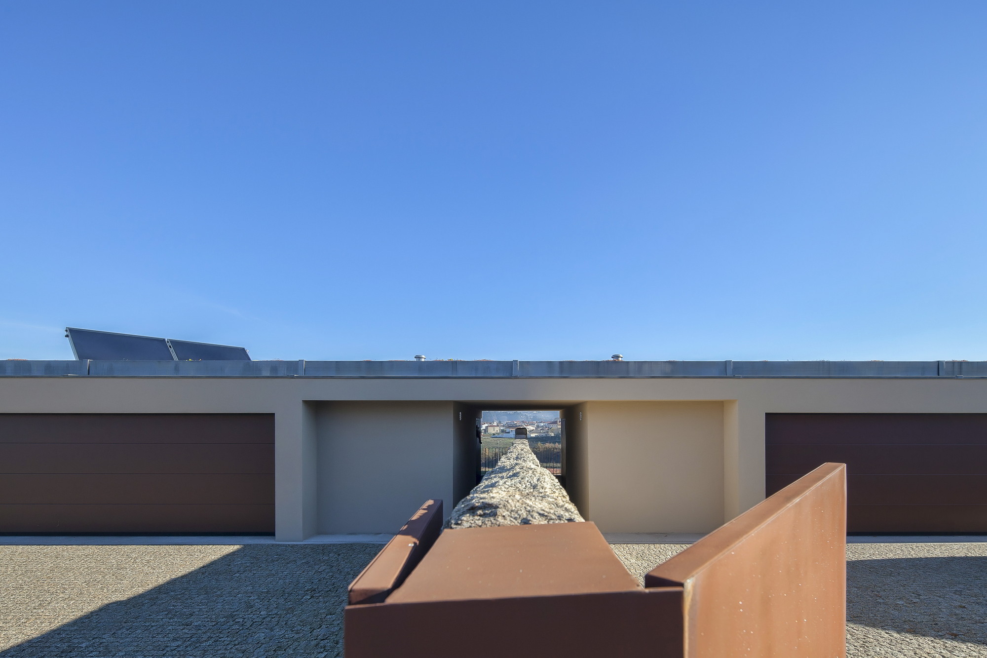 Entrance courtyard seen through weathering steel planters, with a rough stone channel leading to a central opening between two brown garage doors