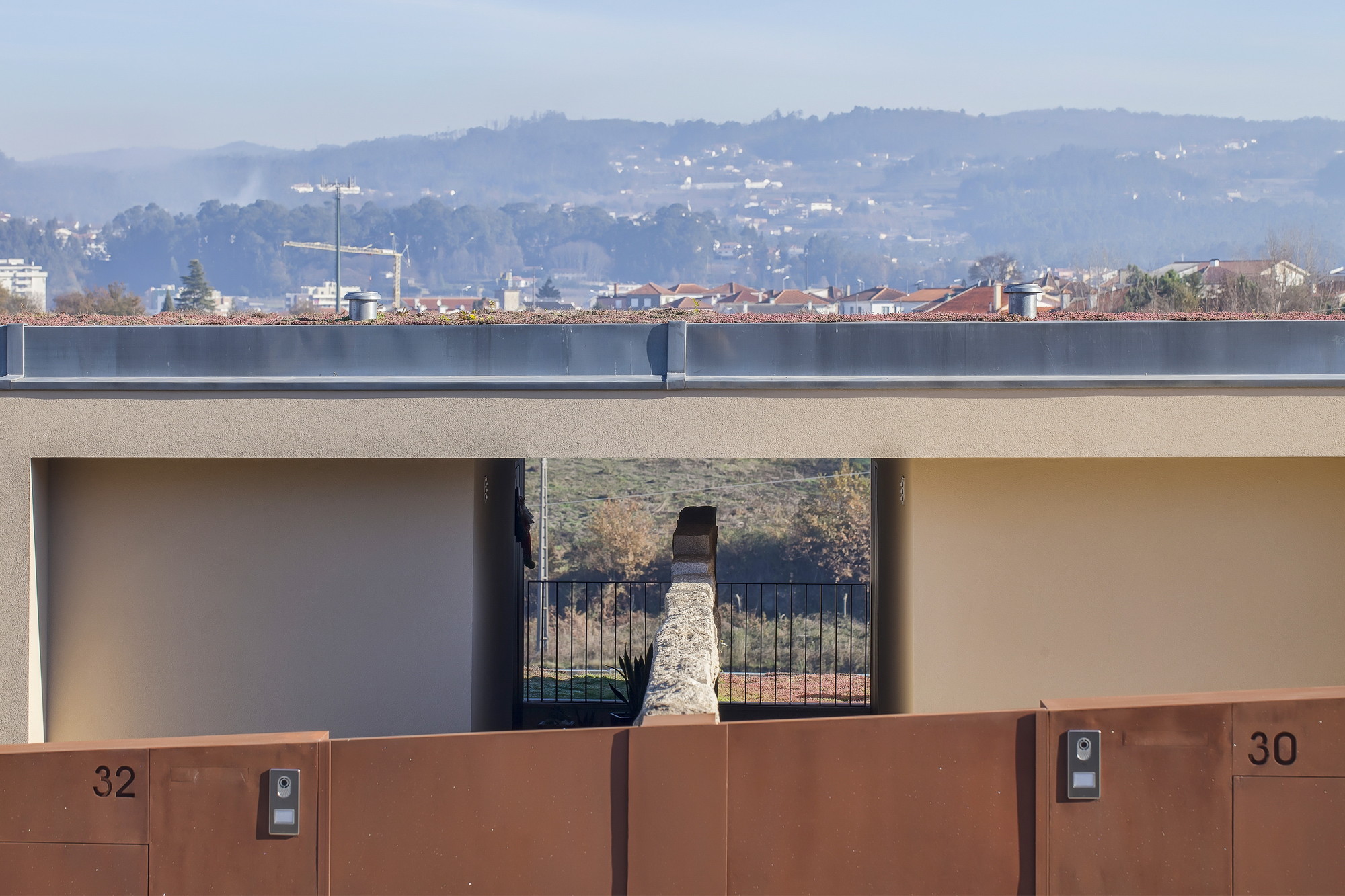 Gate wall with weathering steel panels and house numbers, framing a stone wall and valley view through the open passage under a hazy sky