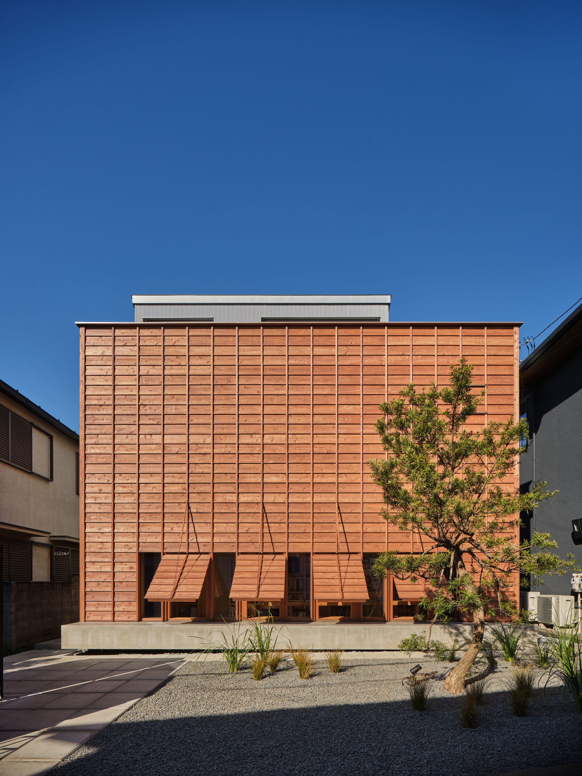 Timber-clad facade with hinged wooden awnings propped open above the ground-floor windows, a pine tree and gravel garden in front under clear blue sky