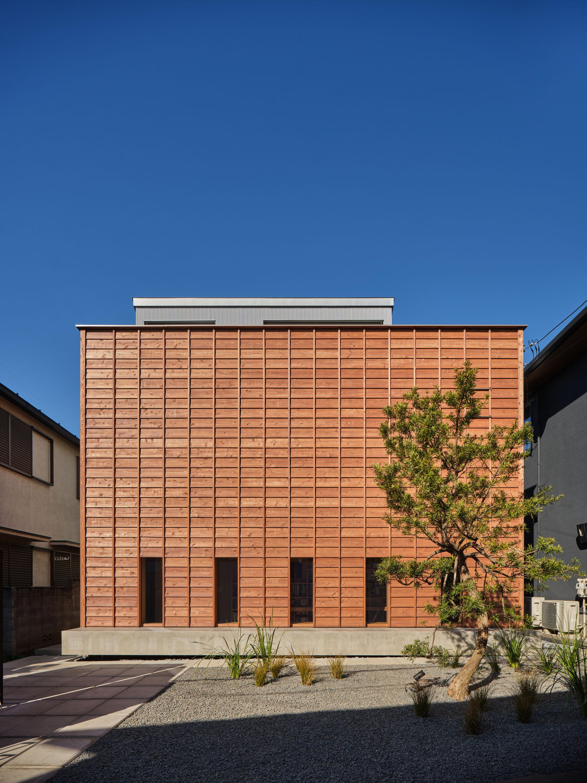 Same timber facade with the awnings closed flat against the cladding, leaving narrow dark window slots above the concrete plinth and a pine tree