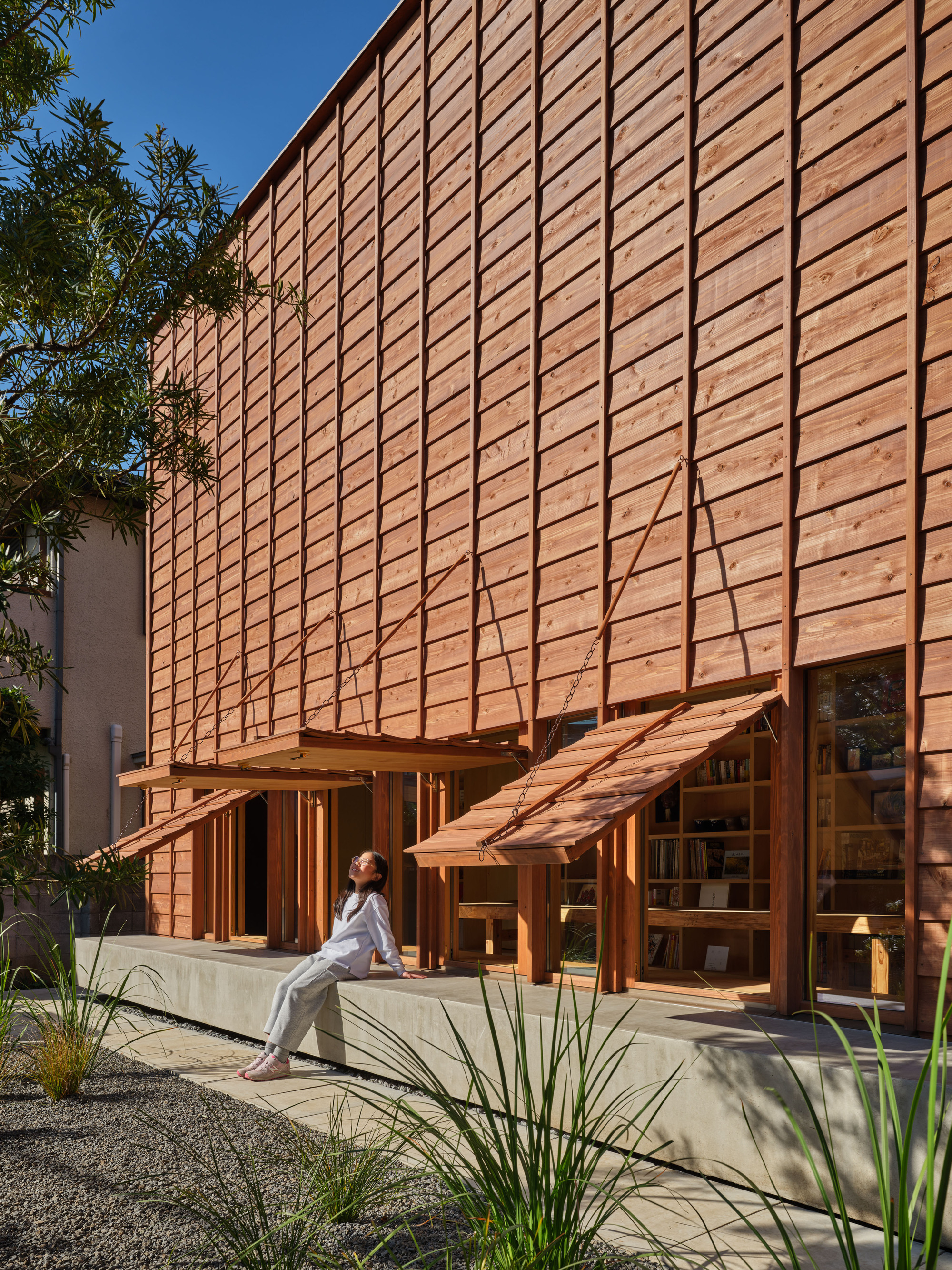 Angled view of the cedar facade with propped awnings, a woman seated on the concrete ledge in front of a bookshelf-lined window
