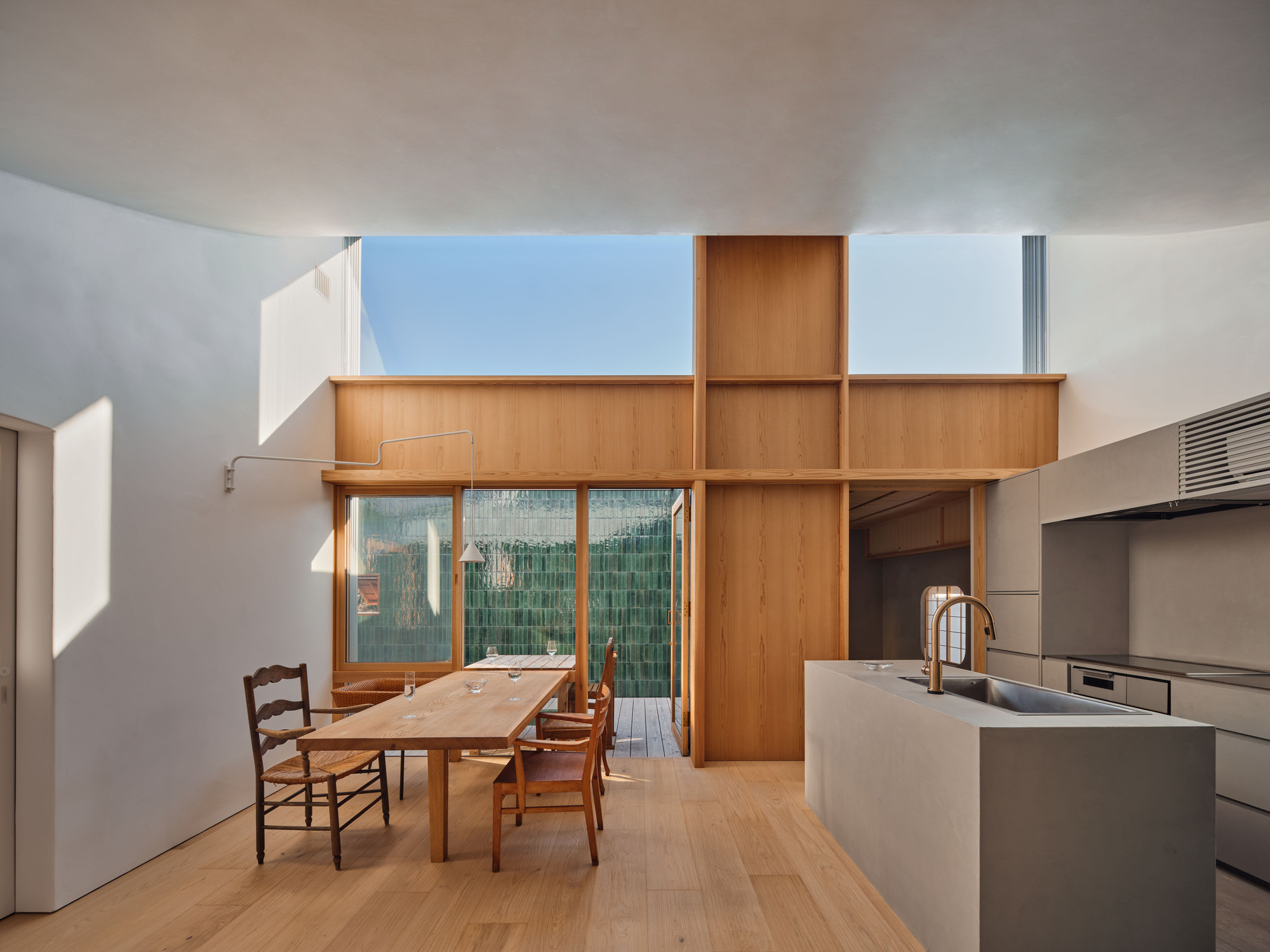 Open kitchen and dining space with oak table, concrete island and timber screen framing a green tiled courtyard wall in afternoon sun