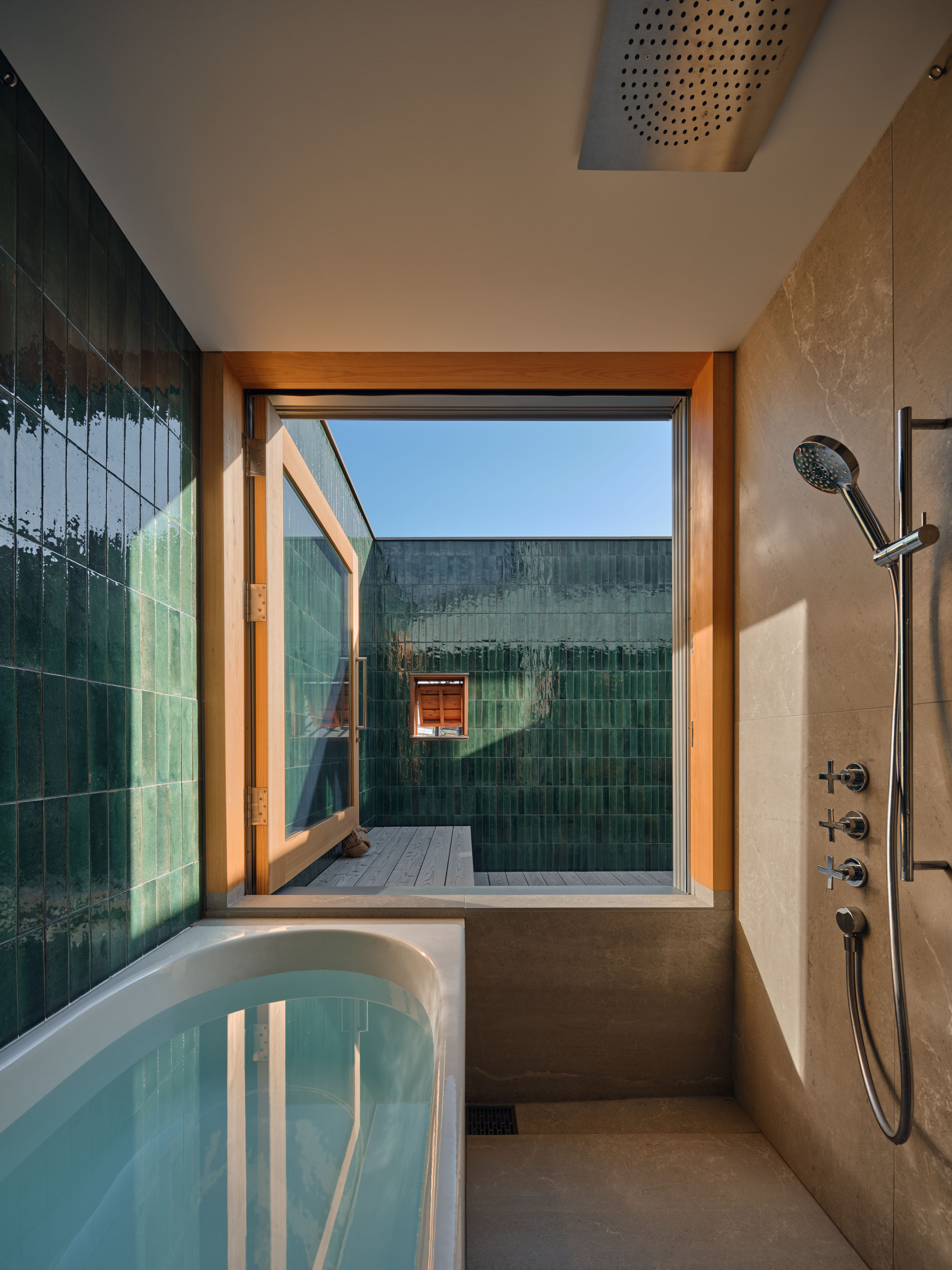 Bathroom with a tub, rain shower head and stone-lined wall, opening through a pivoting window to a green glazed tile courtyard