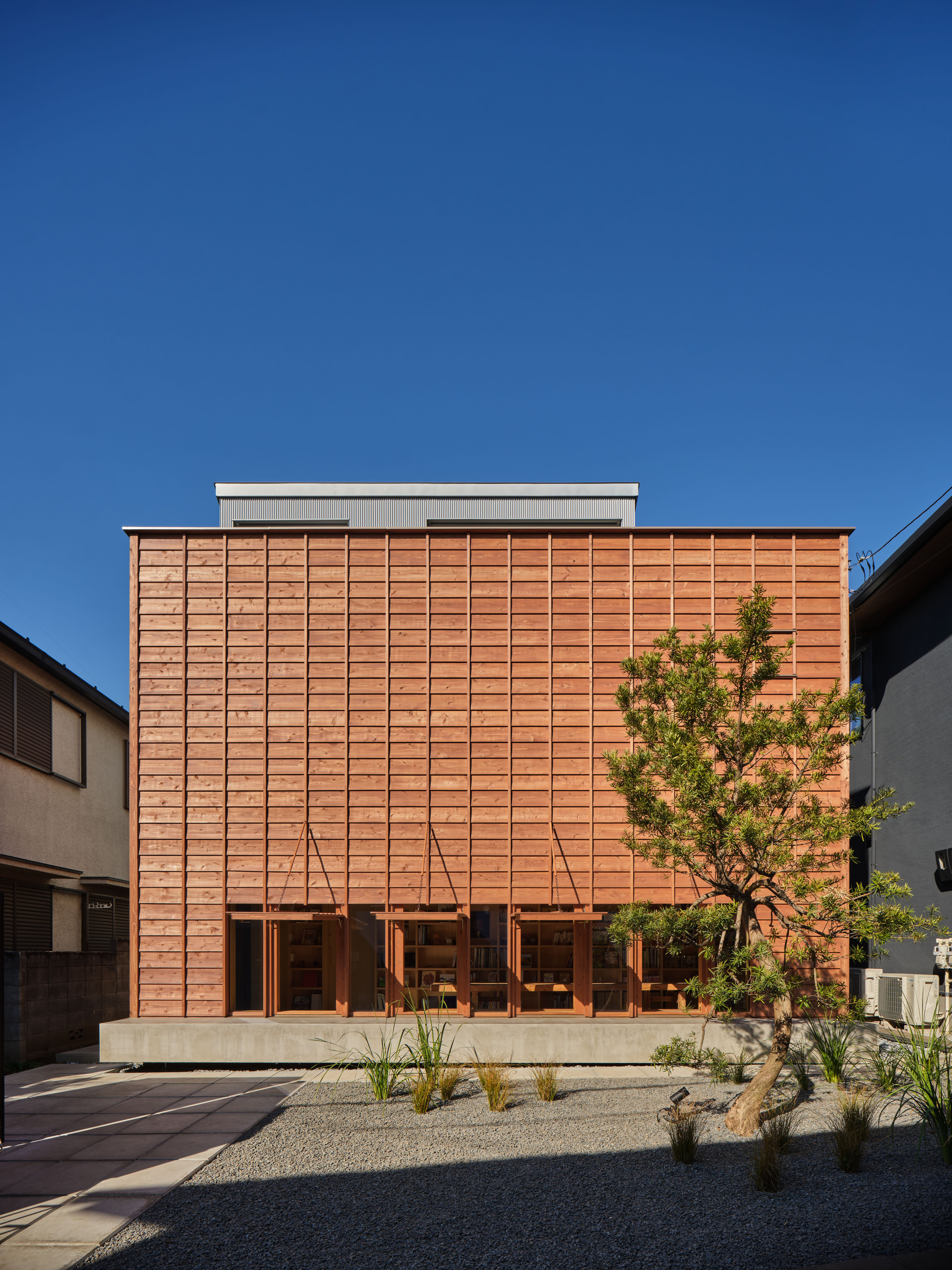 Timber facade with awnings lifted and tied back, showing the lit bookshelves behind the ground-floor glazing beside a pine tree in low sun
