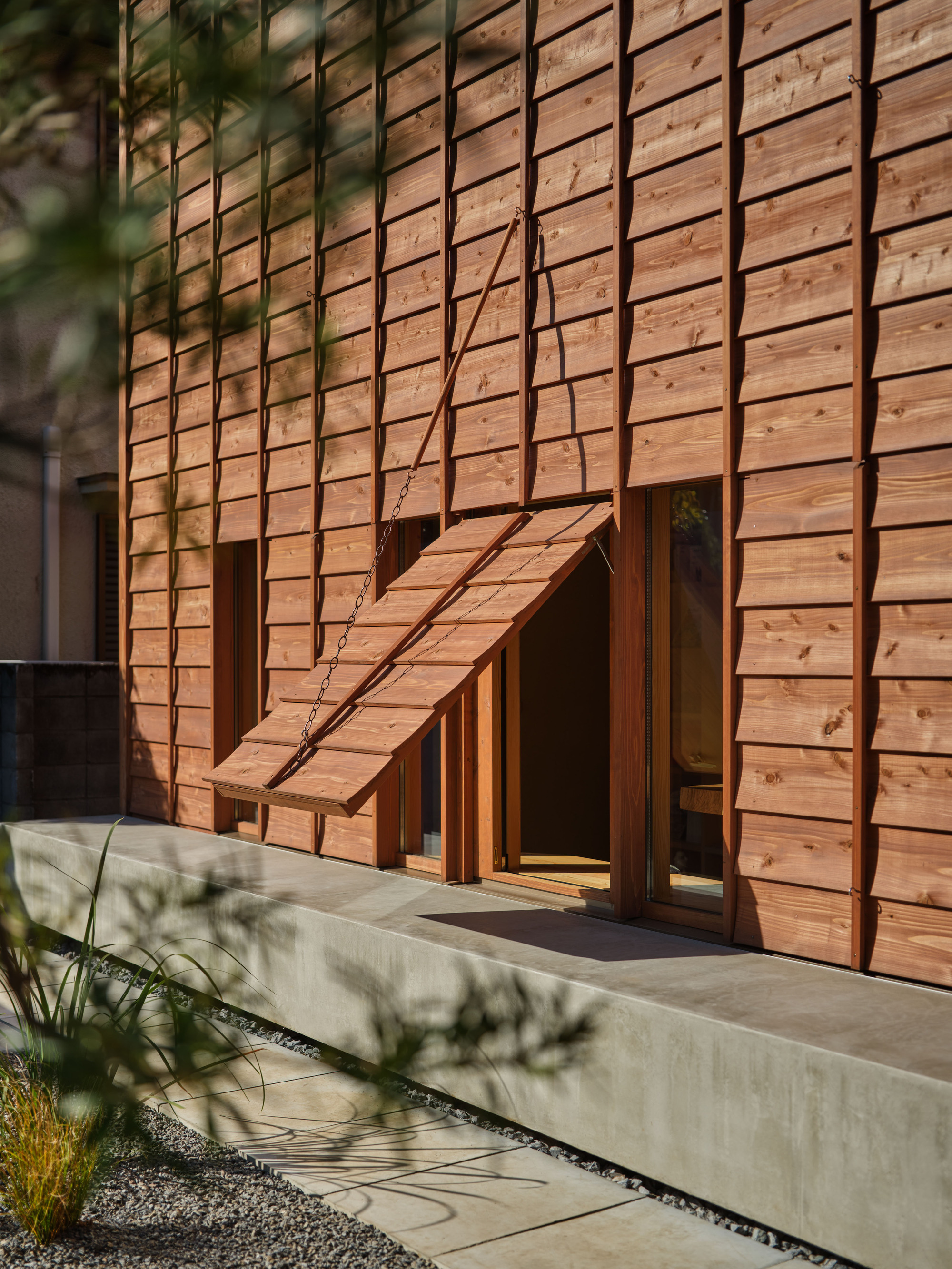 Timber-clad facade with a propped-open wooden shutter held by a chain above a concrete plinth, in dappled afternoon sun
